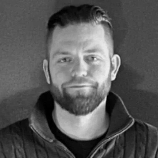 A black and white photo of a man with short hair, beard, and a jacket, smiling slightly.