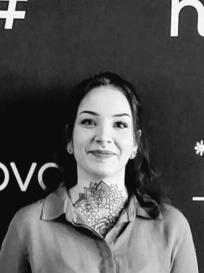 A woman with dark hair, smiling, standing against a dark background with partial text visible. She has a floral tattoo on her neck and is wearing a collared shirt.