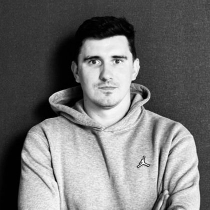 Black and white photo of a young man with dark hair, wearing a light gray hoodie with a small logo, standing against a dark background.