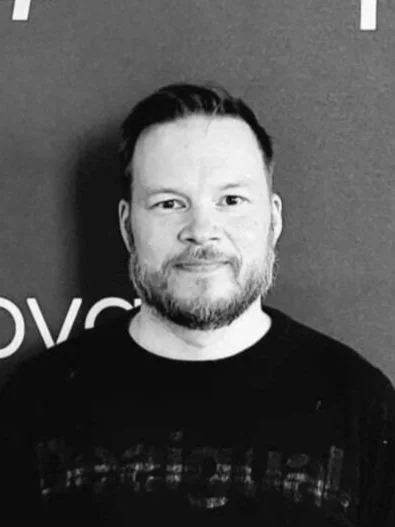 Black and white photo of a man with short dark hair and a beard, smiling, wearing a dark shirt, standing in front of a textured wall.