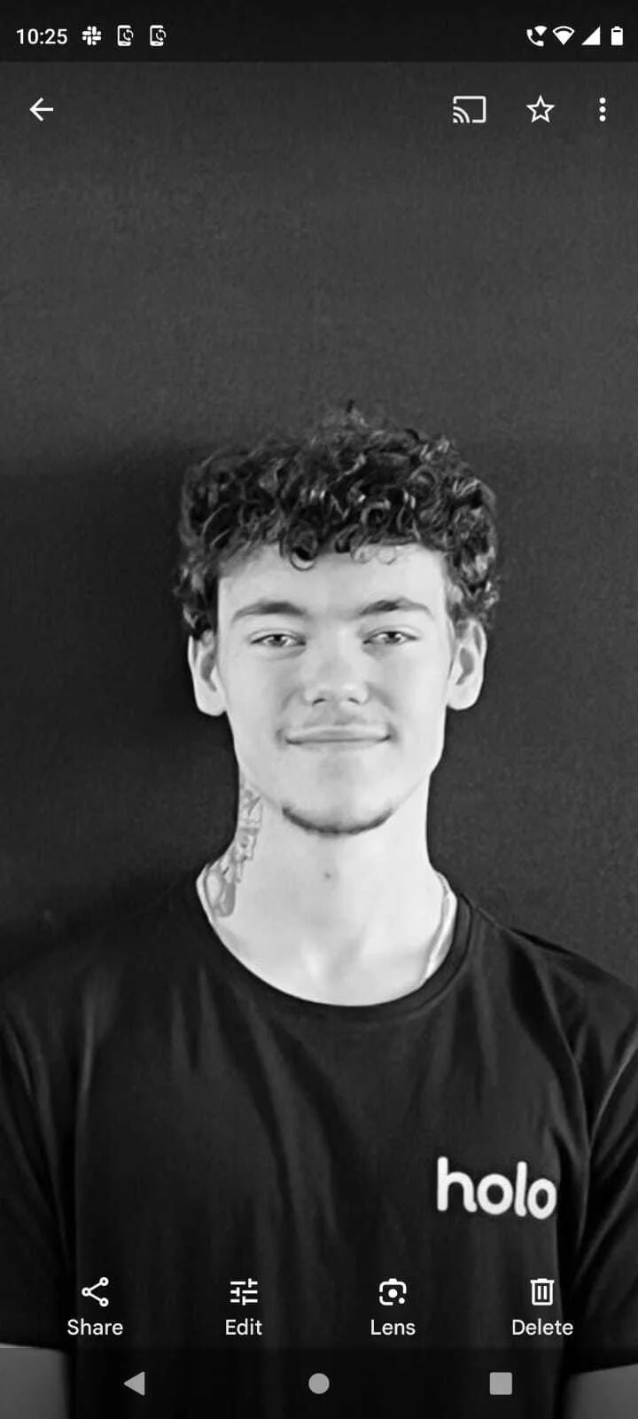 A young man with curly hair, a slight smile, and a tattoo on his neck, wearing a black t-shirt with the word 'holo' printed on it, taking a selfie against a dark background.