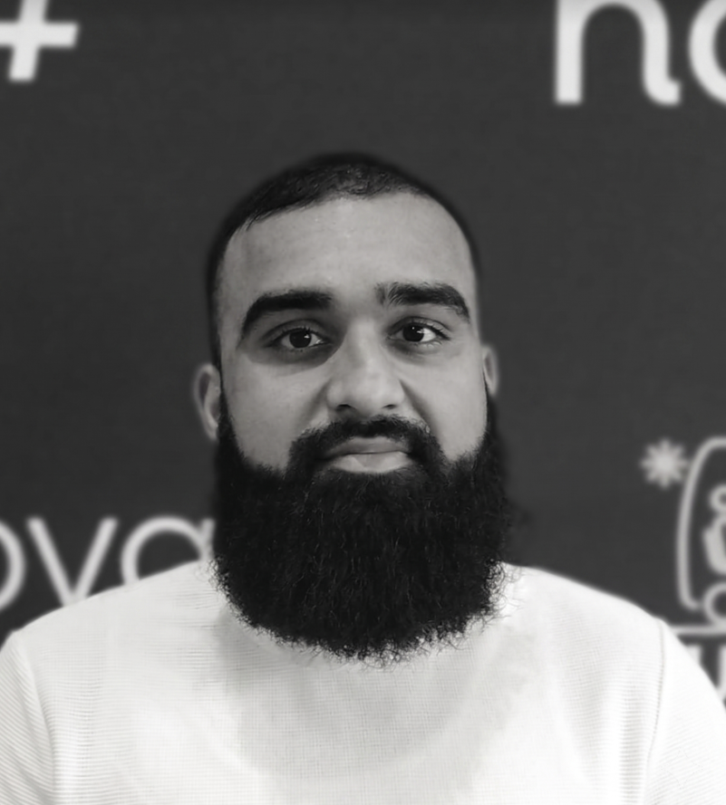A man with a full beard, mustache, and short dark hair, looking directly at the camera with a neutral expression. He is wearing a light-colored shirt, and the background appears to be a dark wall with partial text or logos.