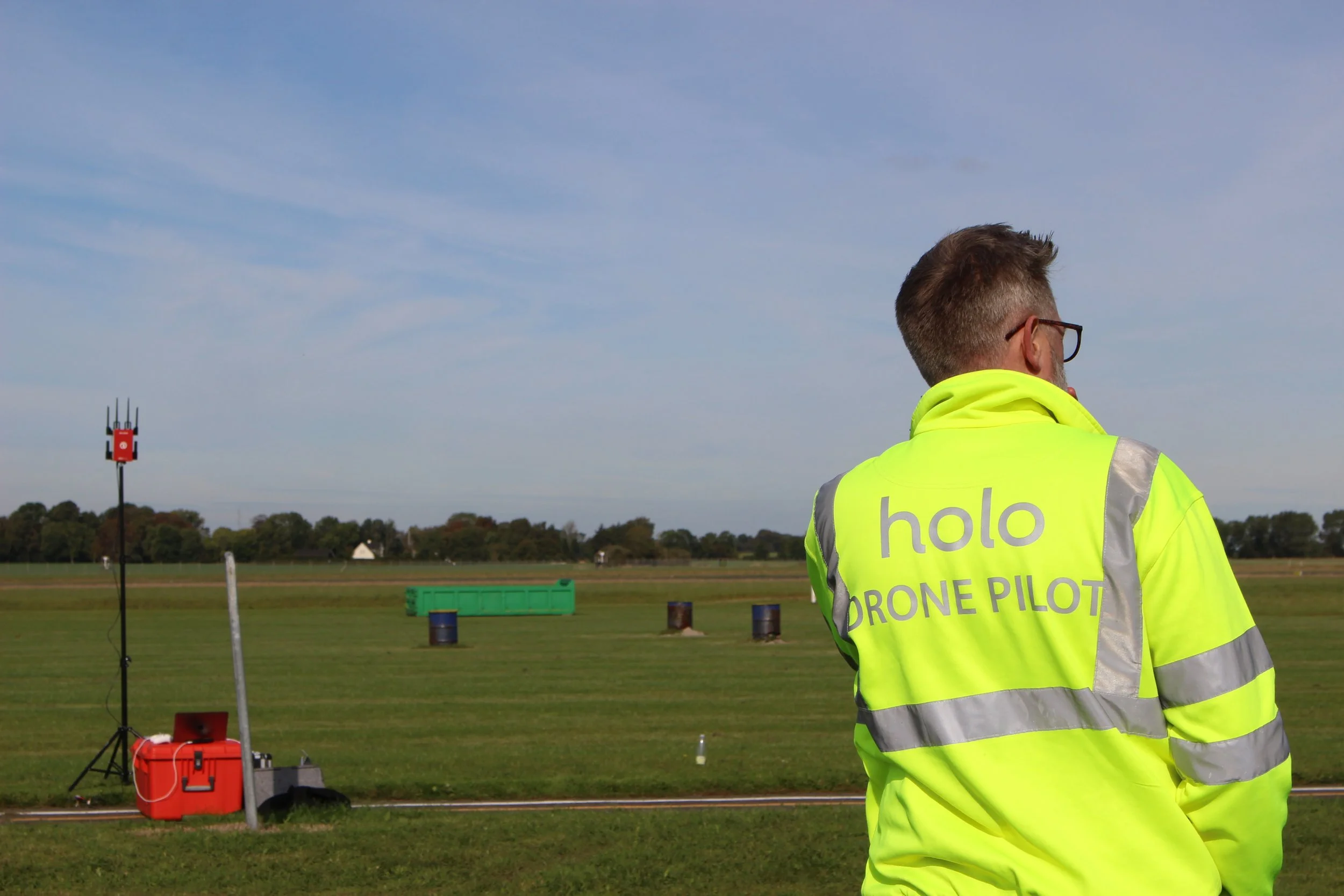A drone pilot wearing a high-visibility yellow jacket with 'holo DRONE PILOT' on the back, standing outside on a field, with a weather station or antenna tower and equipment in the background under a blue sky.