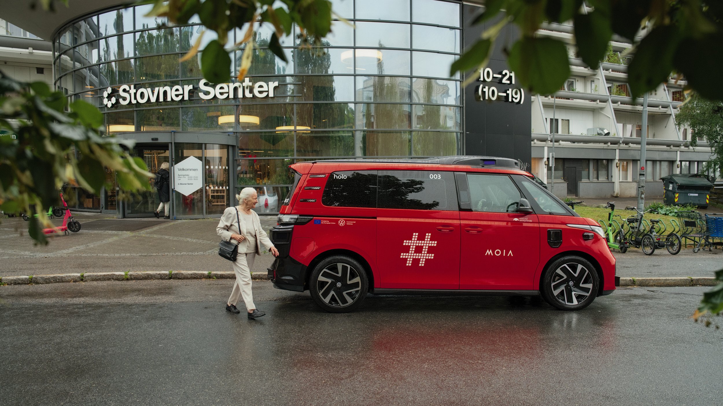 An elderly woman walking past a red autonomous vehicle in front of the Stovner Senter shopping mall, with people, bicycles, and greenery visible around.