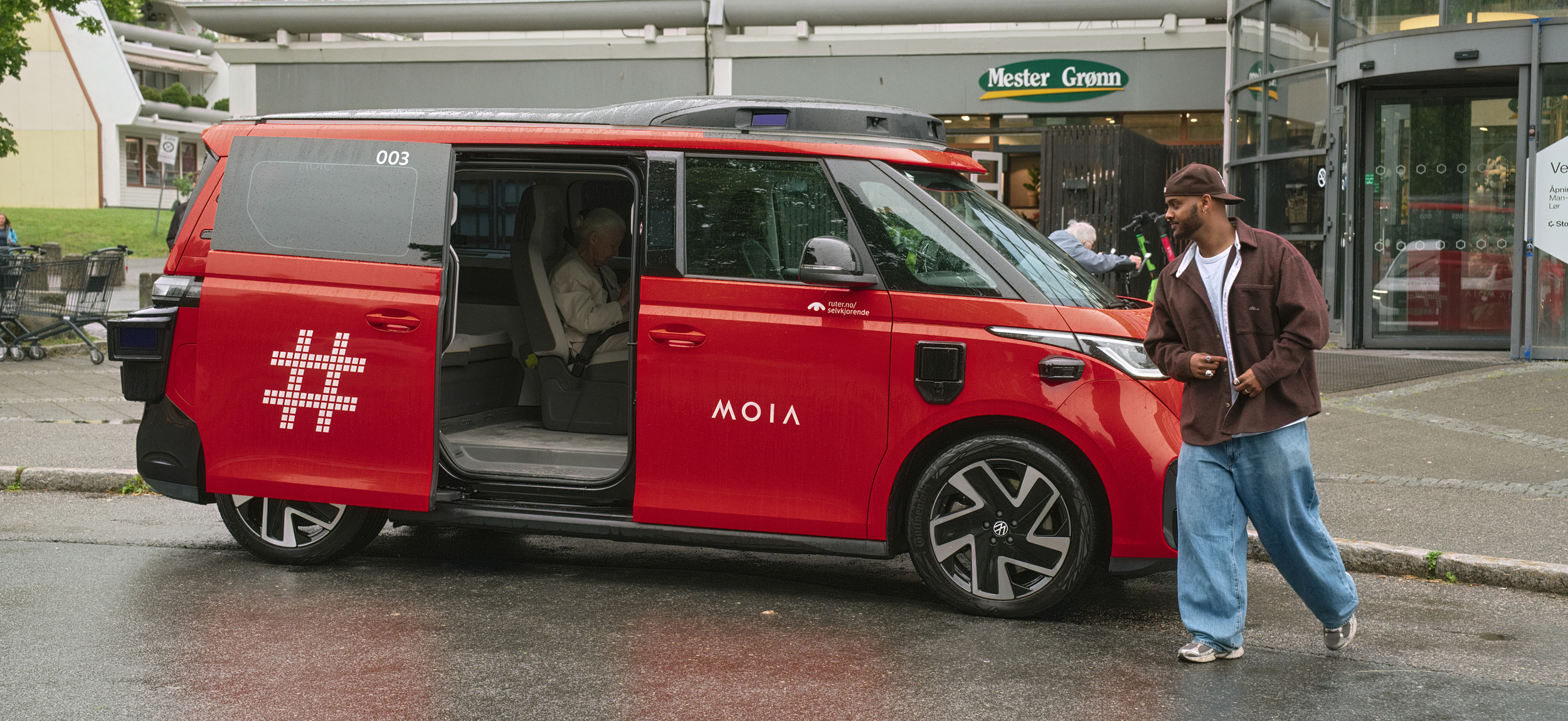 A red autonomous shuttle vehicle with open sliding door parked outside a building. Inside, an elderly person is seated, while a young man in a brown jacket and blue jeans stands outside, looking at his phone.