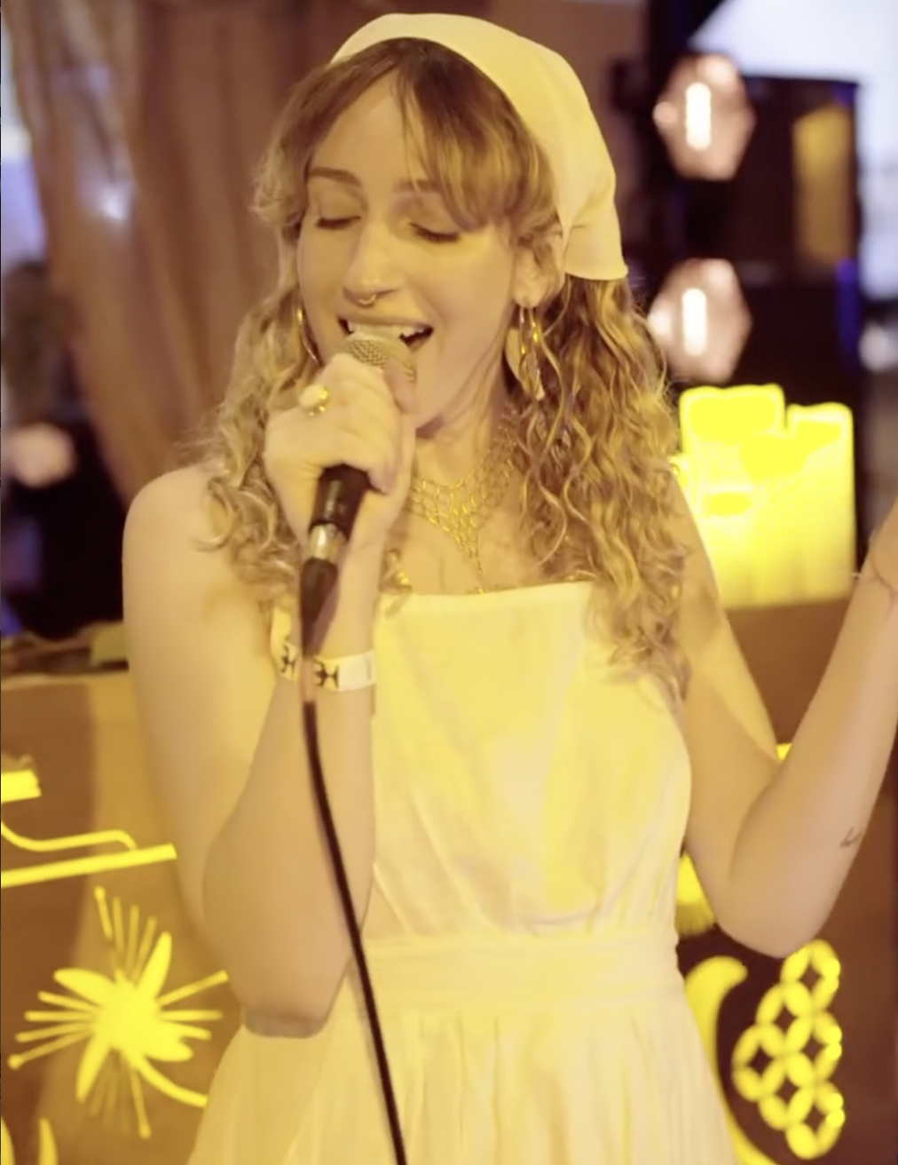 A woman with curly hair, wearing a yellow dress and headscarf, is singing into a microphone at a bar or venue illuminated with yellow and warm lighting.