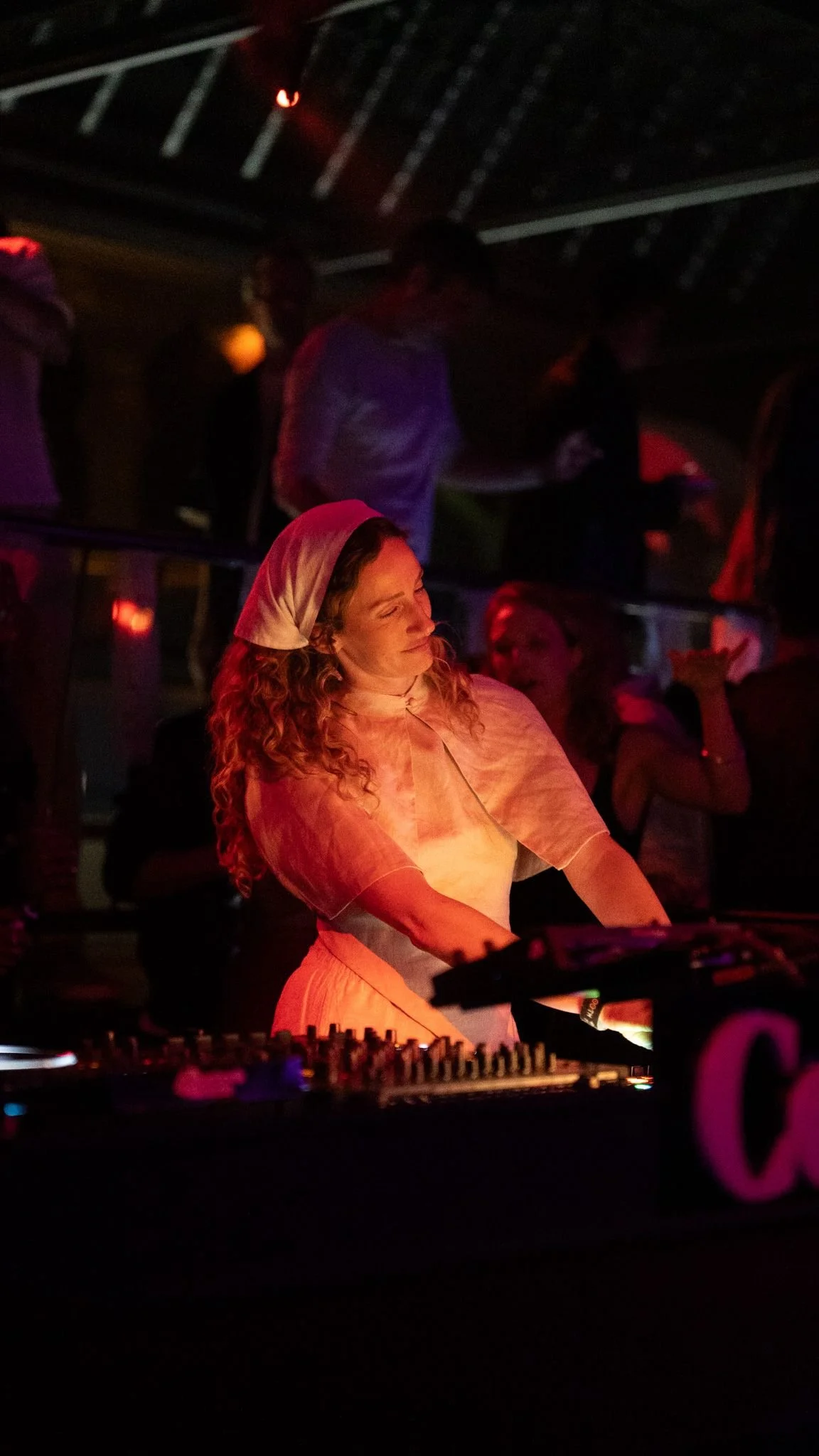 A woman with curly hair wearing a white headscarf and white dress behind DJ equipment at a dimly lit party, with other people in the background.