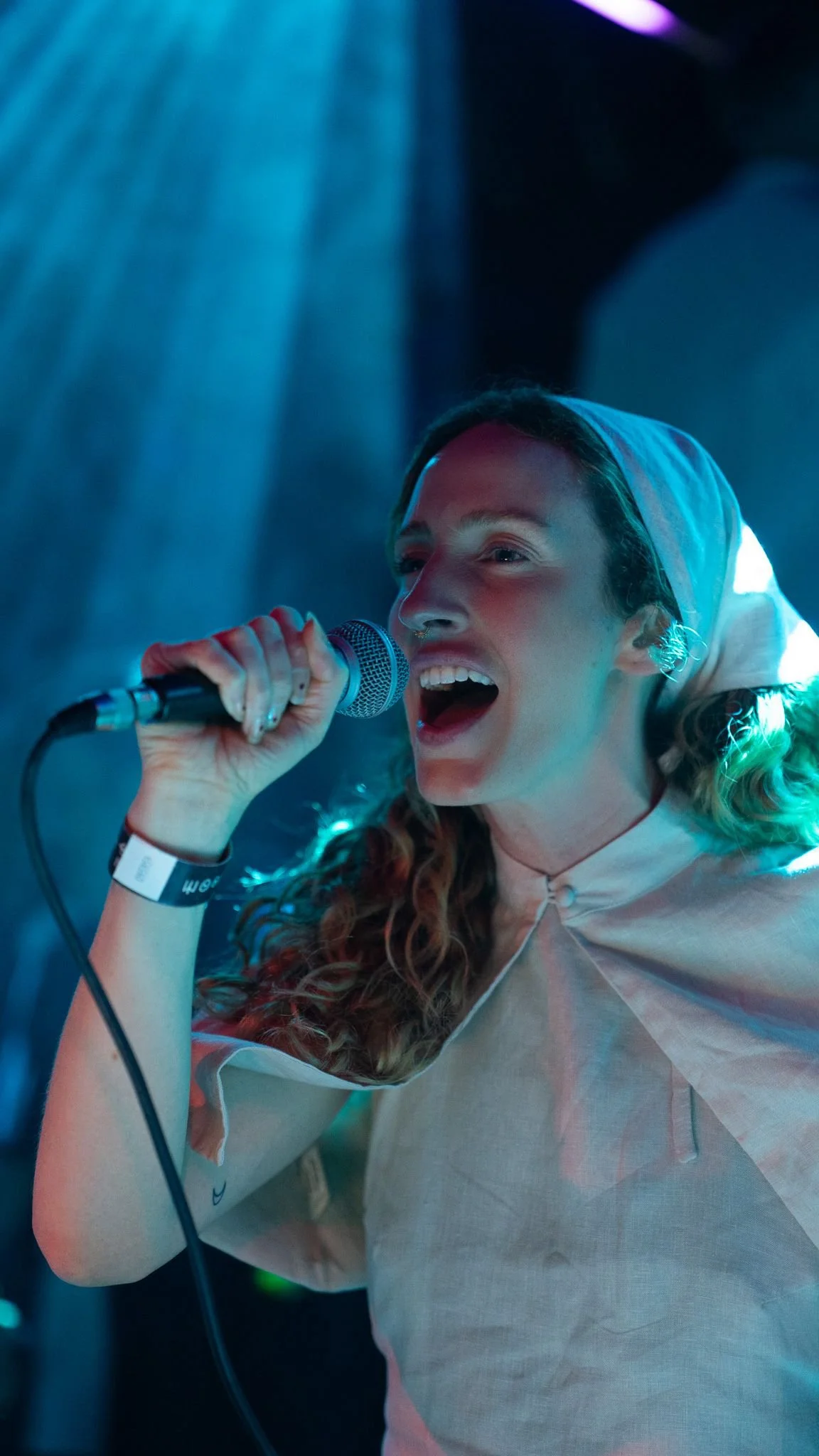 A young woman singing into a microphone on stage, wearing a hooded garment with stage lighting creating a colorful ambiance.