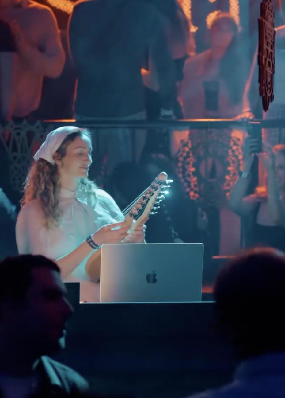 A young woman with curly hair, wearing a white headscarf and a white top, is playing a xylophone at a music event, with a laptop and people in the background.
