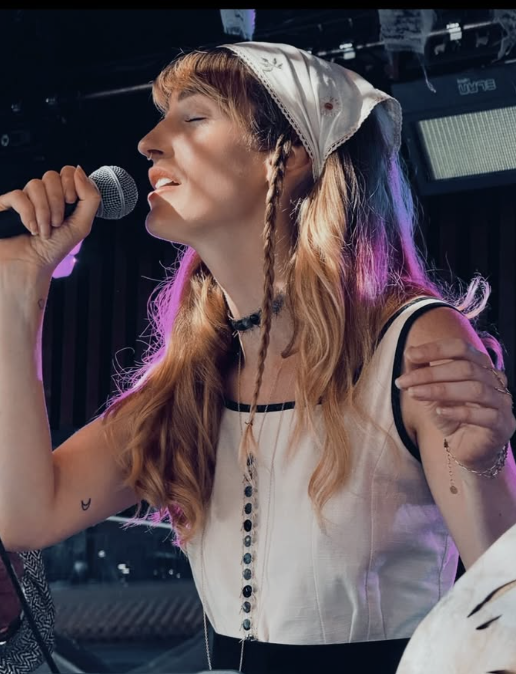 A woman with long, wavy blonde hair with purple streaks, wearing a white bandana, singing into a microphone with her eyes closed and an expressive facial expression, on stage with colorful lighting.