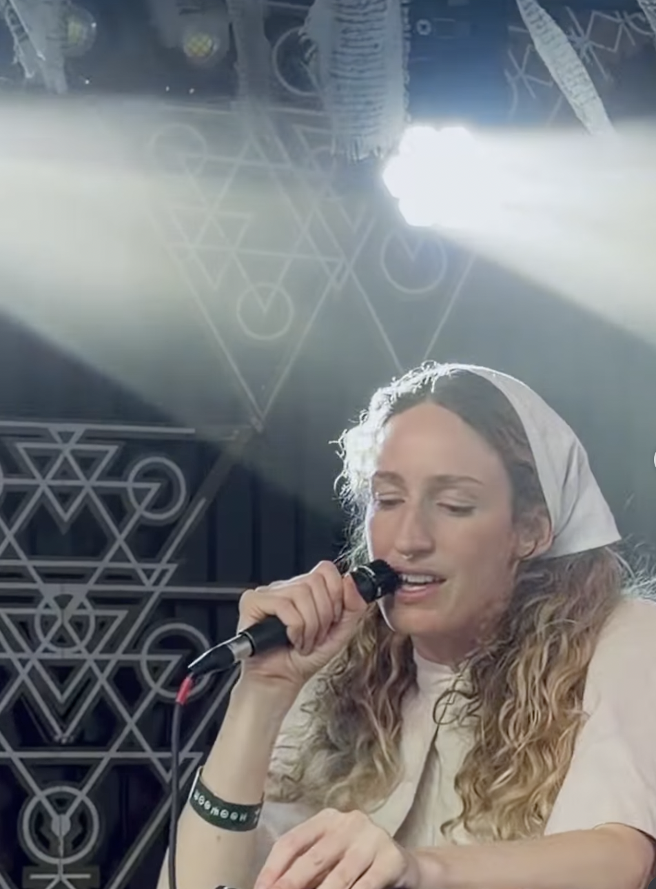 A woman with curly blonde hair wearing a white headscarf, holding a microphone and speaking or singing, with a geometric patterned wall and symbols in the background, illuminated by a bright light.