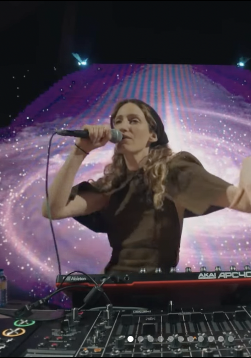 Female DJ with long curly hair performing with a microphone and DJ equipment in front of a vibrant galaxy-themed backdrop.