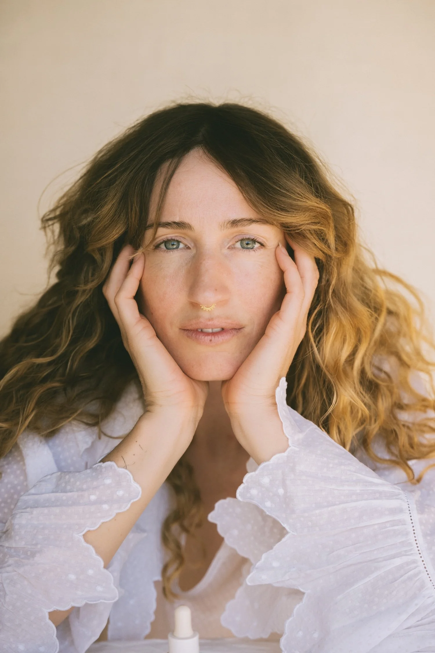 A woman with long, curly hair holding her face with her hands, looking at the camera, wearing a white lace top, sitting at a table with a small white bottle in front of her, in a neutral-toned room.