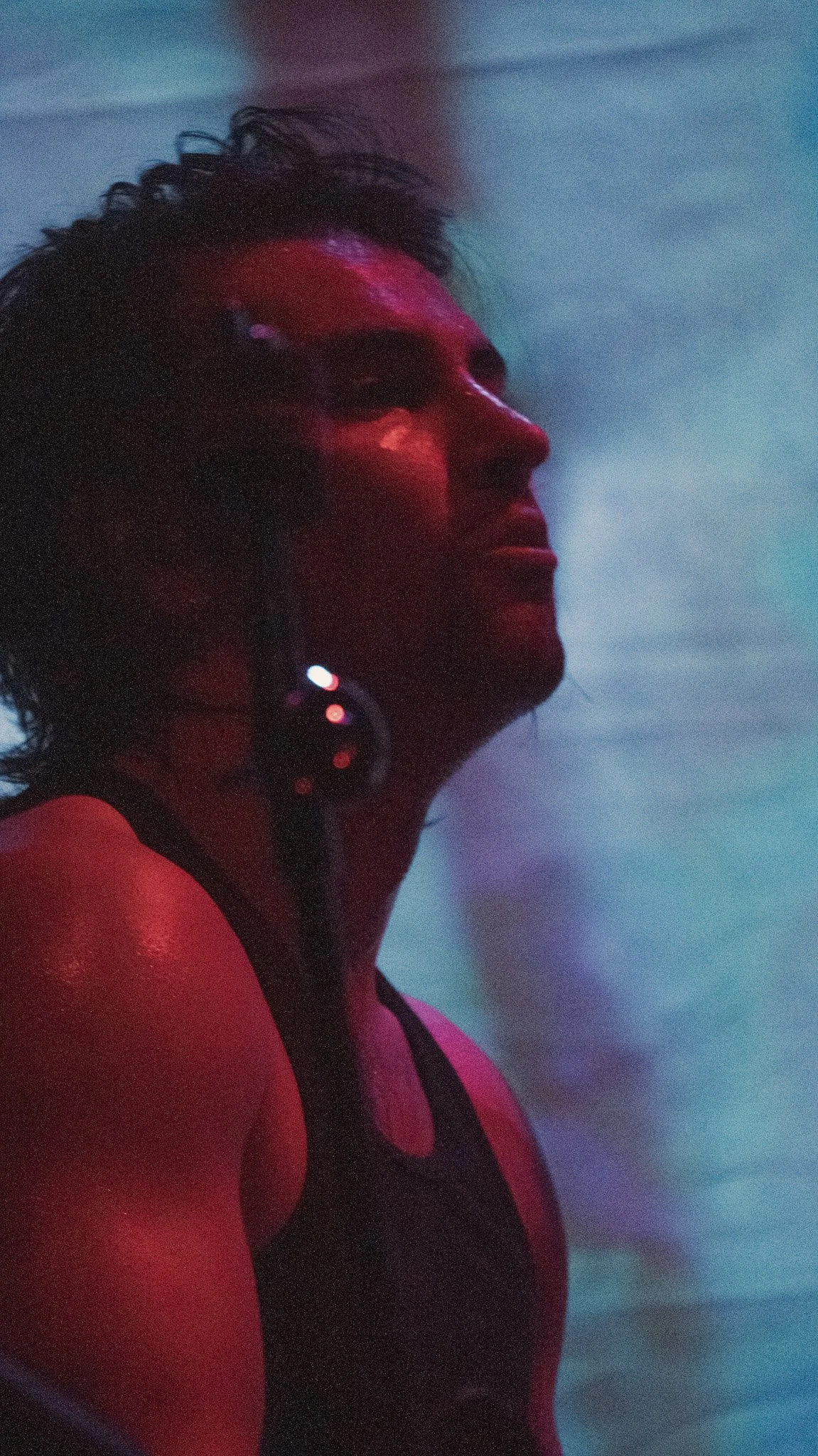 A woman with short curly hair in water, illuminated by pink and blue lighting, wearing a black swimsuit and earrings.