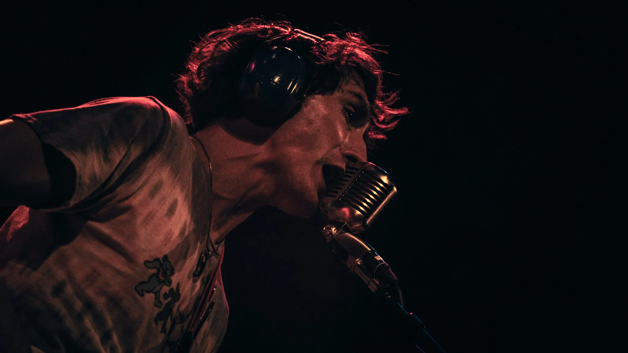 A person singing into a vintage microphone, wearing large headphones and a Mickey Mouse T-shirt, with red hair illuminated by red lighting against a dark background.