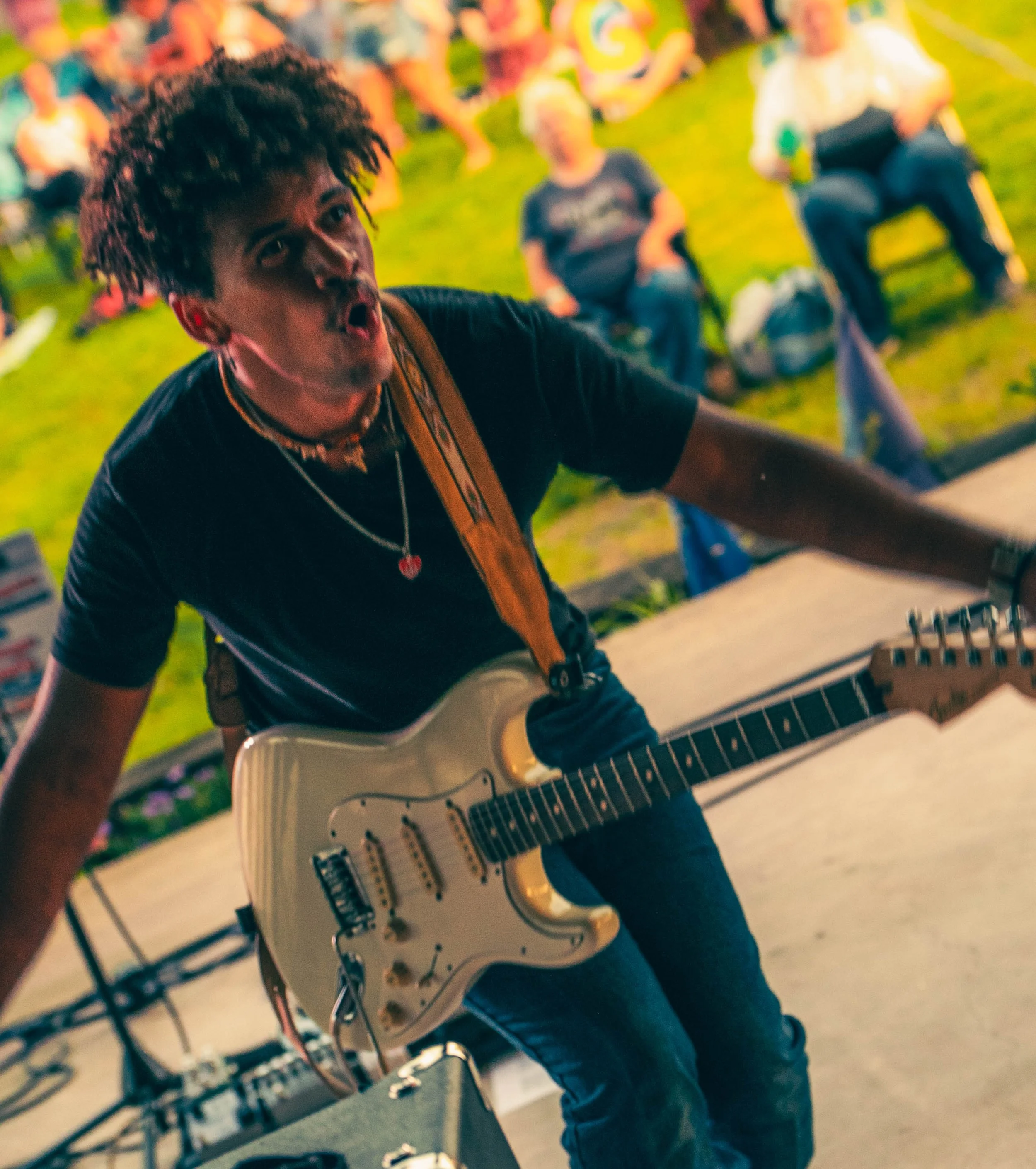 A young man with curly hair playing an electric guitar in a crowded indoor setting, possibly at a music event or party.