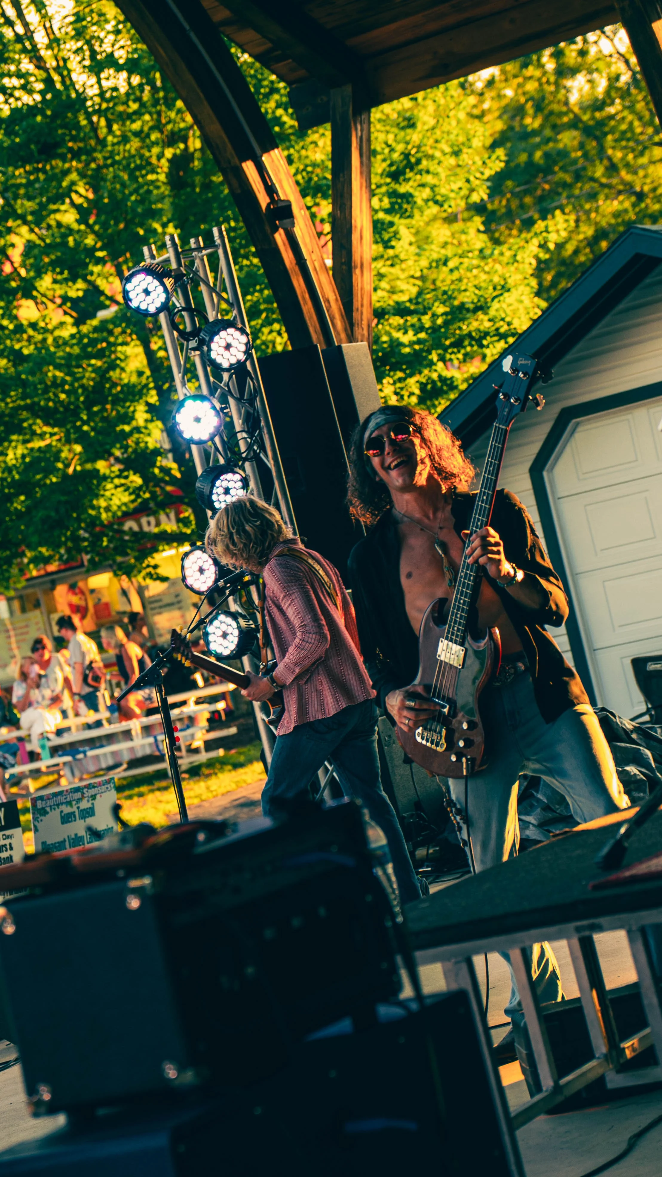 Musicians perform on an outdoor stage during a festival, with green trees in the background, stage lighting, and a crowd in the distance.