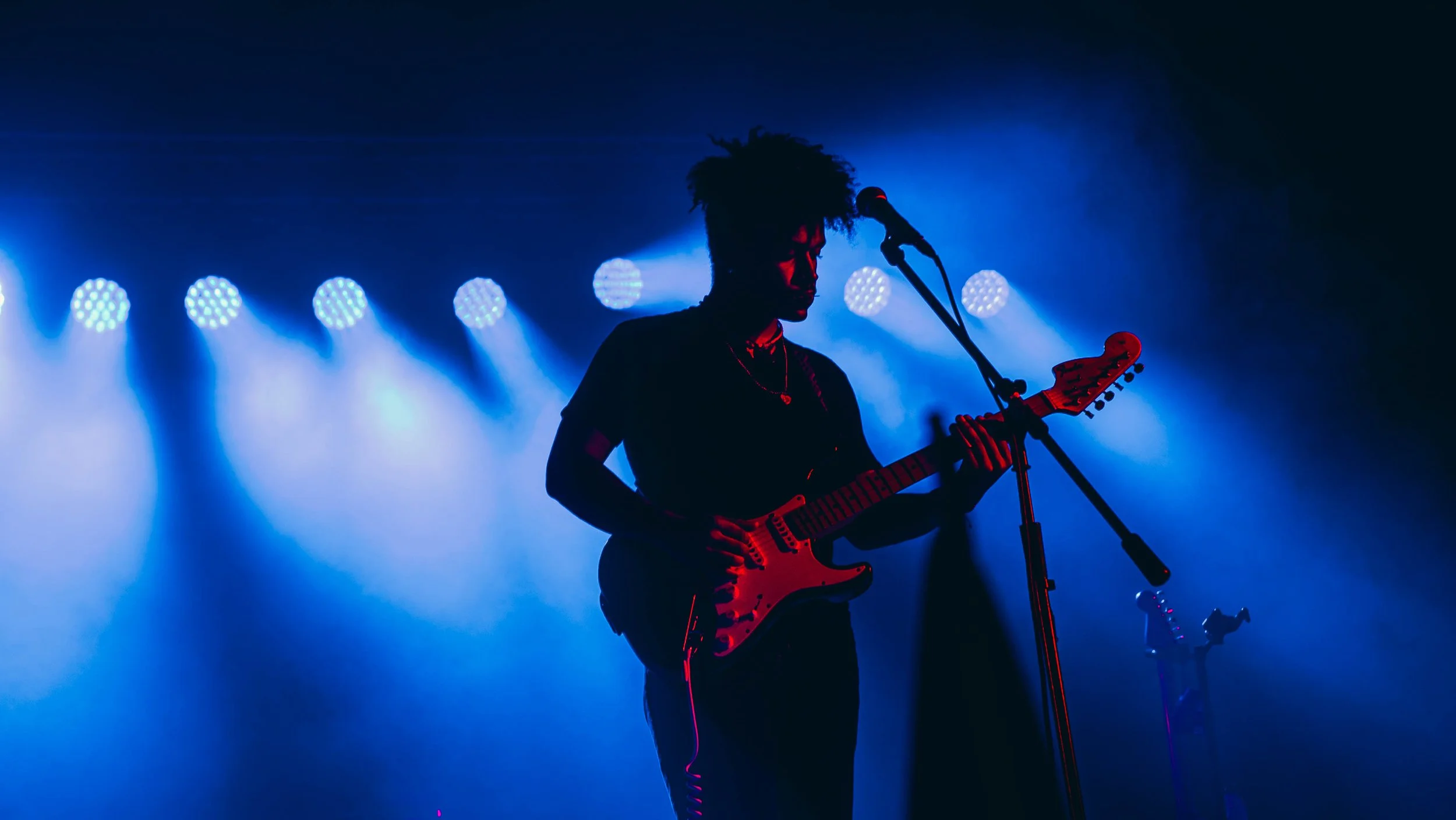 A musician playing an electric guitar on stage, silhouetted against blue stage lights, with a microphone stand in front and fog creating a moody atmosphere.