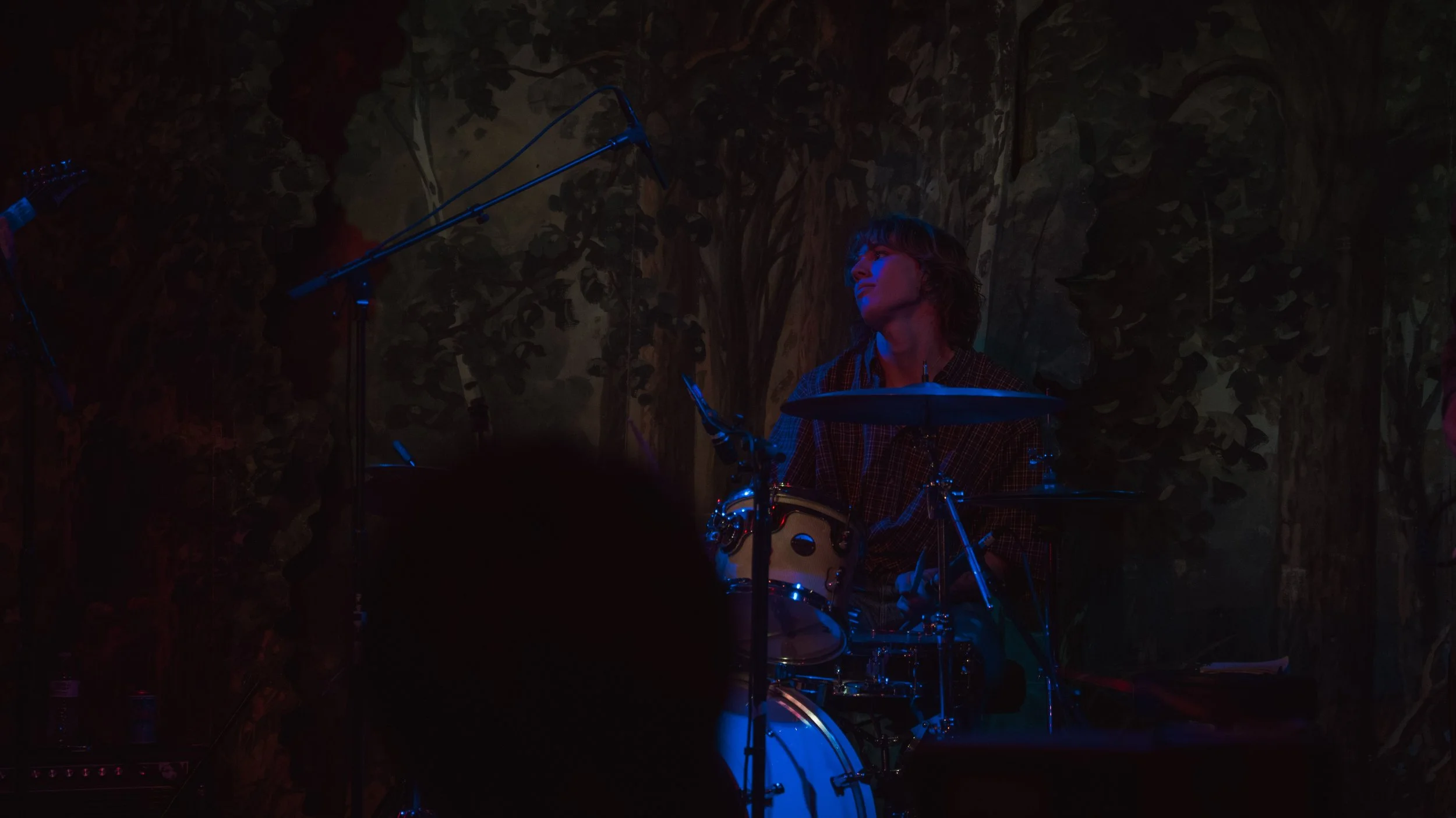 A person is playing drums on stage with a mural background, under blue and red lighting.