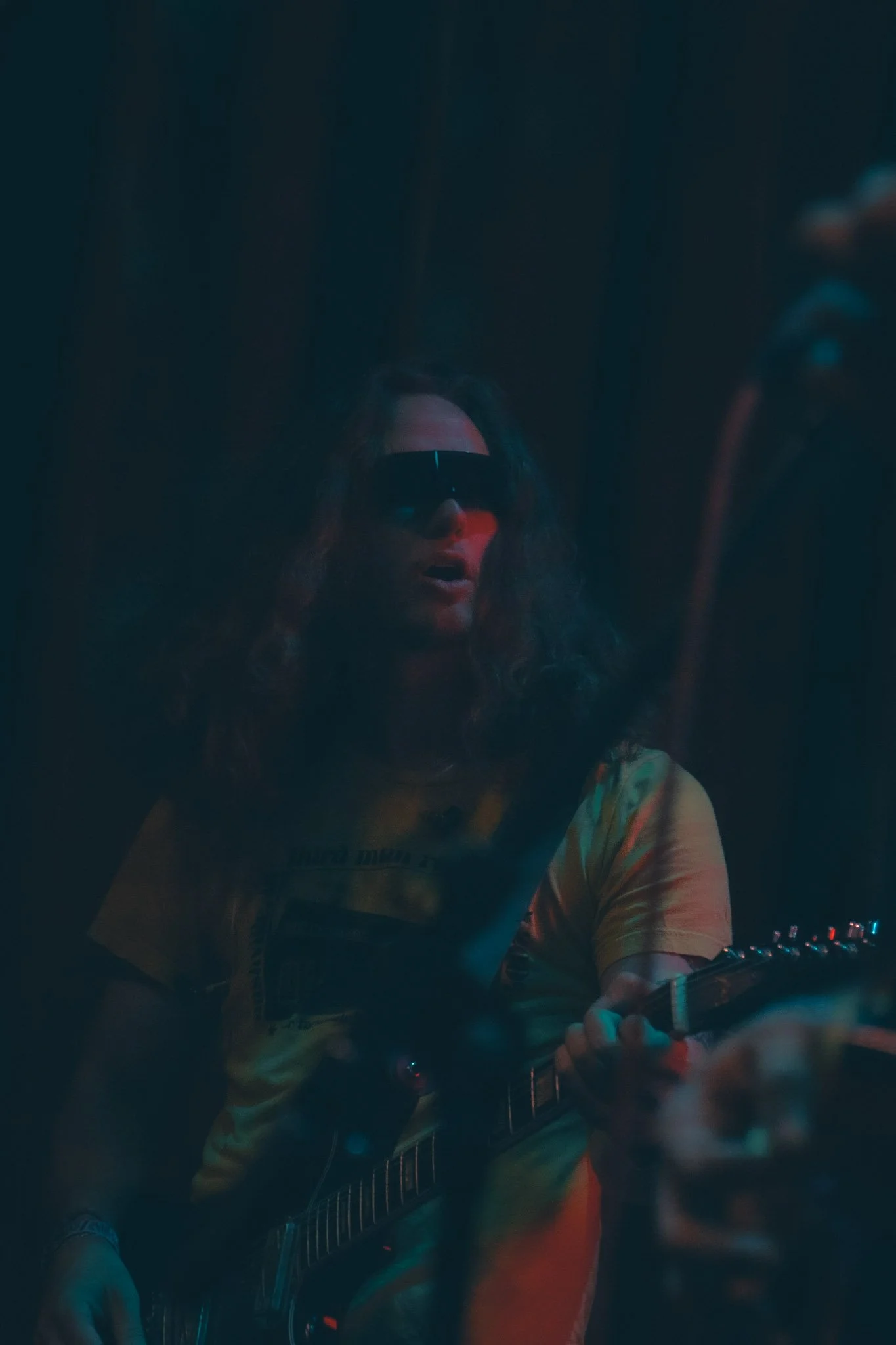 A woman wearing sunglasses and a colorful shirt is playing an electric guitar in a dark, dimly lit room.