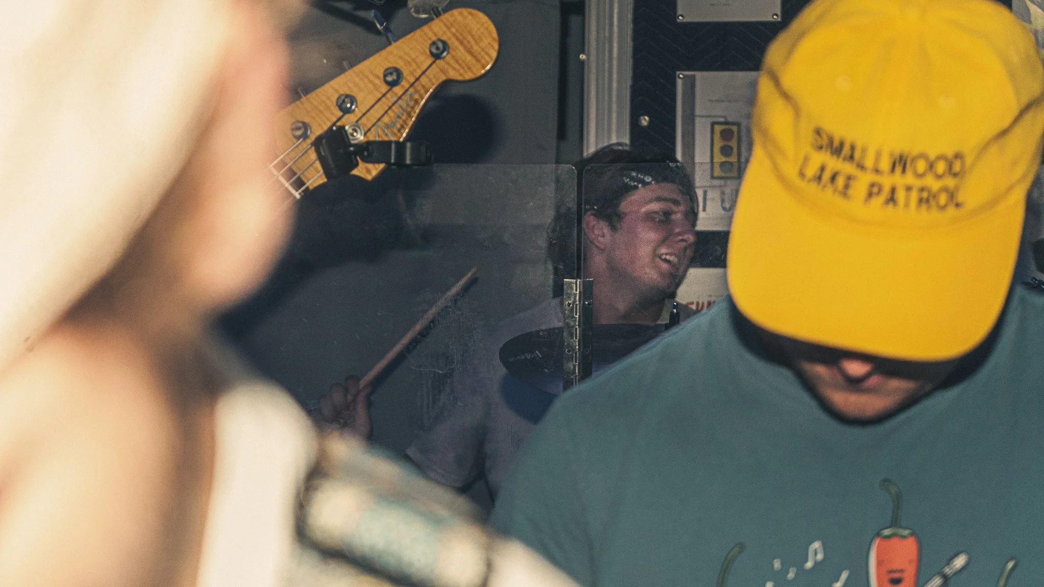 A person with wet hair, smiling, looking at a reflection in a mirror. The person is surrounded by others, one wearing a yellow cap that reads 'SMALLWOOD LAKE PATROL' and another with a drumstick in hand. A guitar is visible in the background.