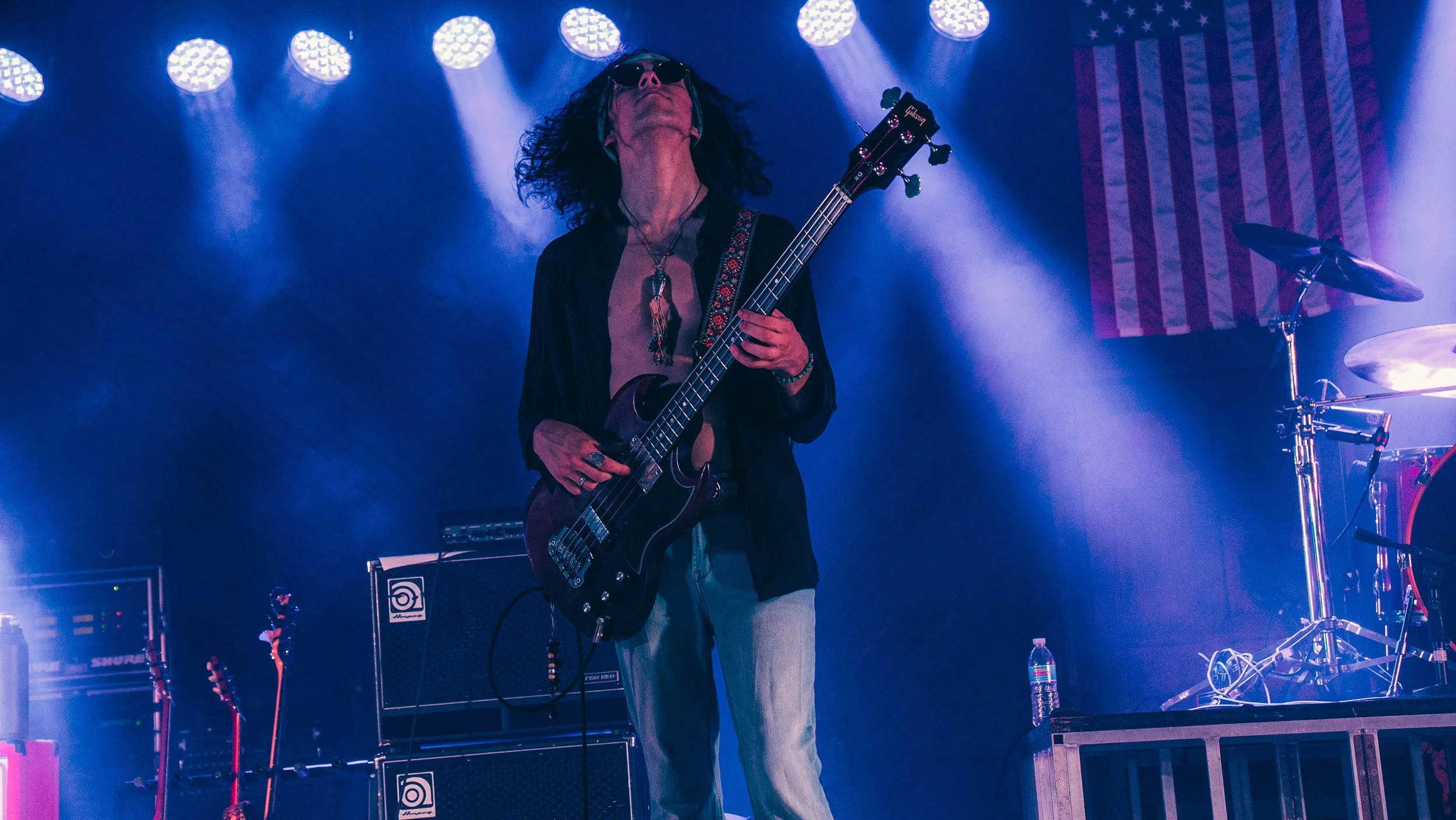Musician playing electric guitar on stage with blue and purple lighting, American flag backdrop, wearing sunglasses and jewelry.