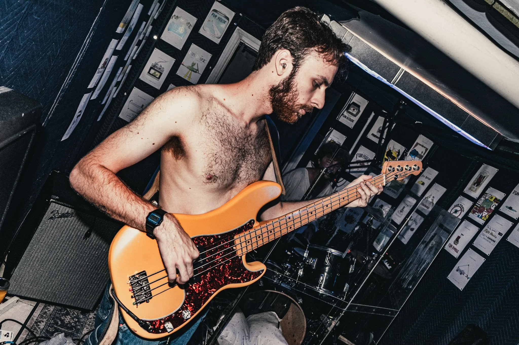 A shirtless man with a beard playing an orange electric bass guitar in a music venue or small stage setting.