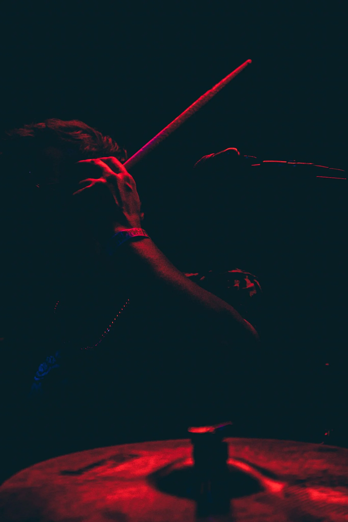 A person appears to be playing drums in a dark setting illuminated by red and blue lights, with only parts of their body and drumsticks visible.