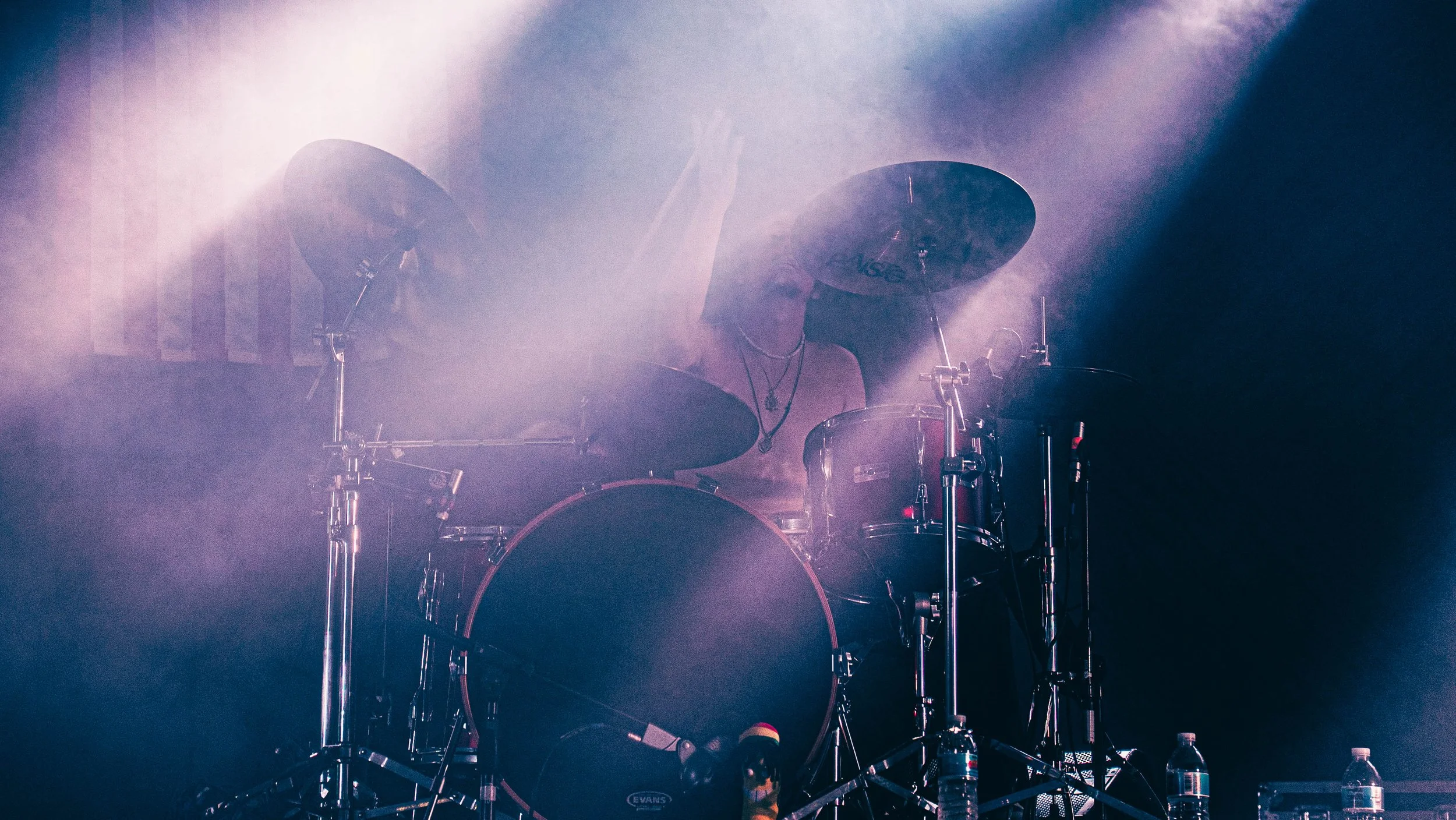 A person playing a drum set on stage with fog and bright lights.