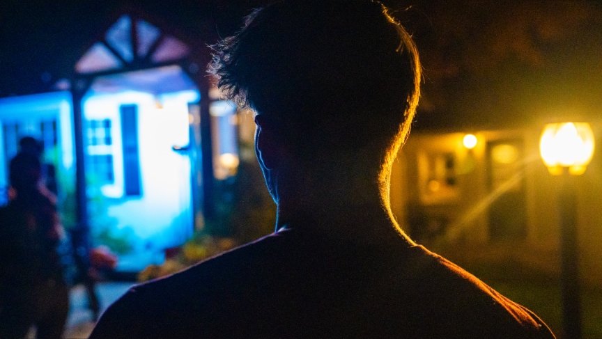Silhouette of a person with short hair standing outside at night, illuminated by warm yellow and cool blue lights.