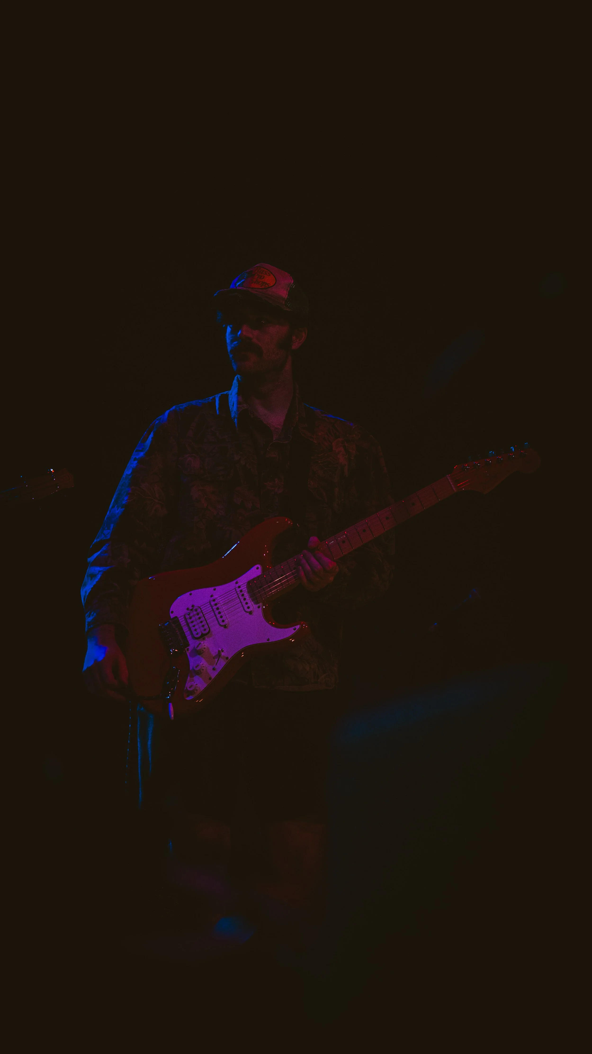 A man wearing a patterned shirt and a baseball cap playing an electric guitar on stage under dark lighting with red and blue lights.
