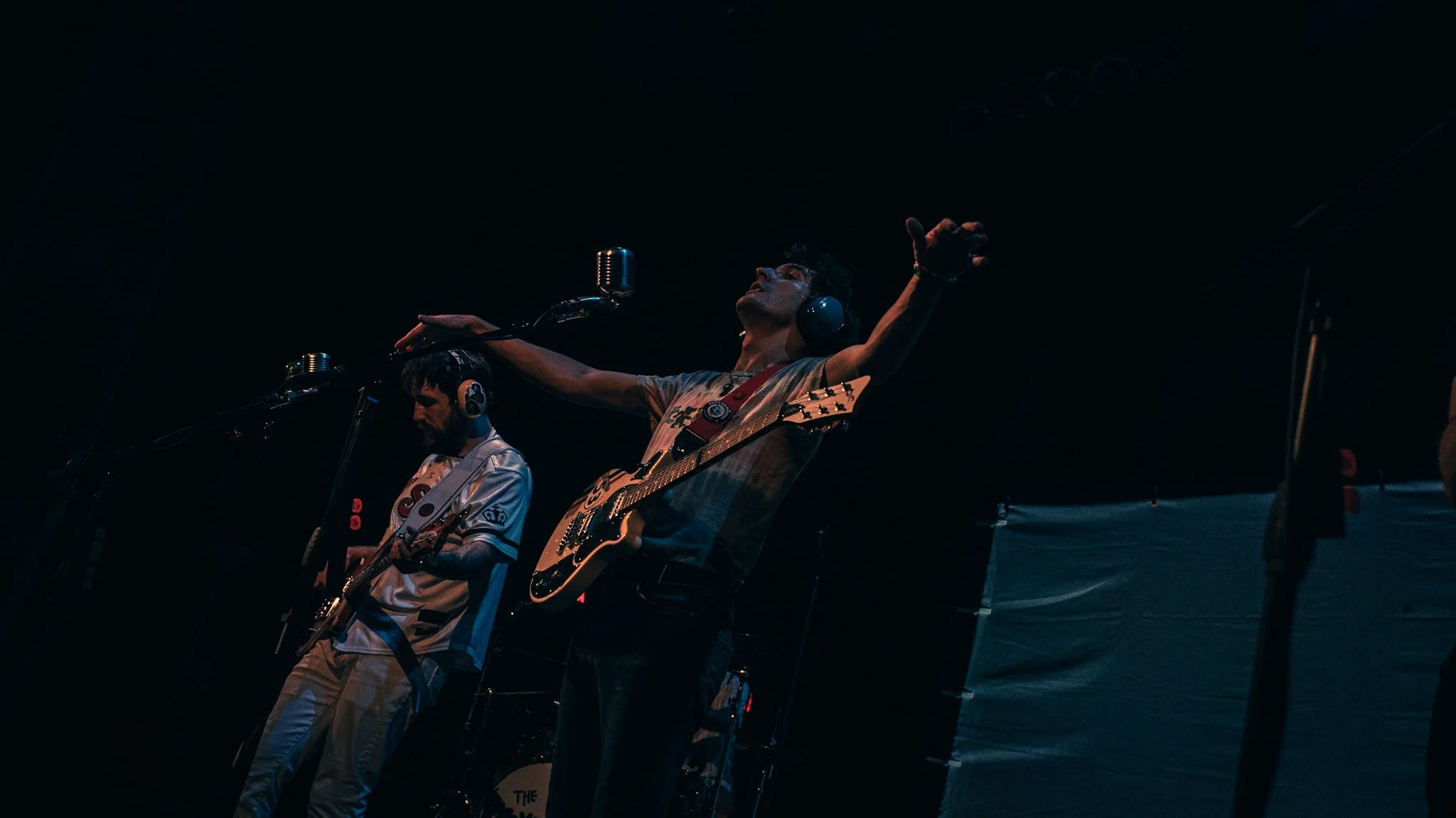 Two male musicians performing on stage with guitars, wearing headphones, in a dark setting with minimal lighting.
