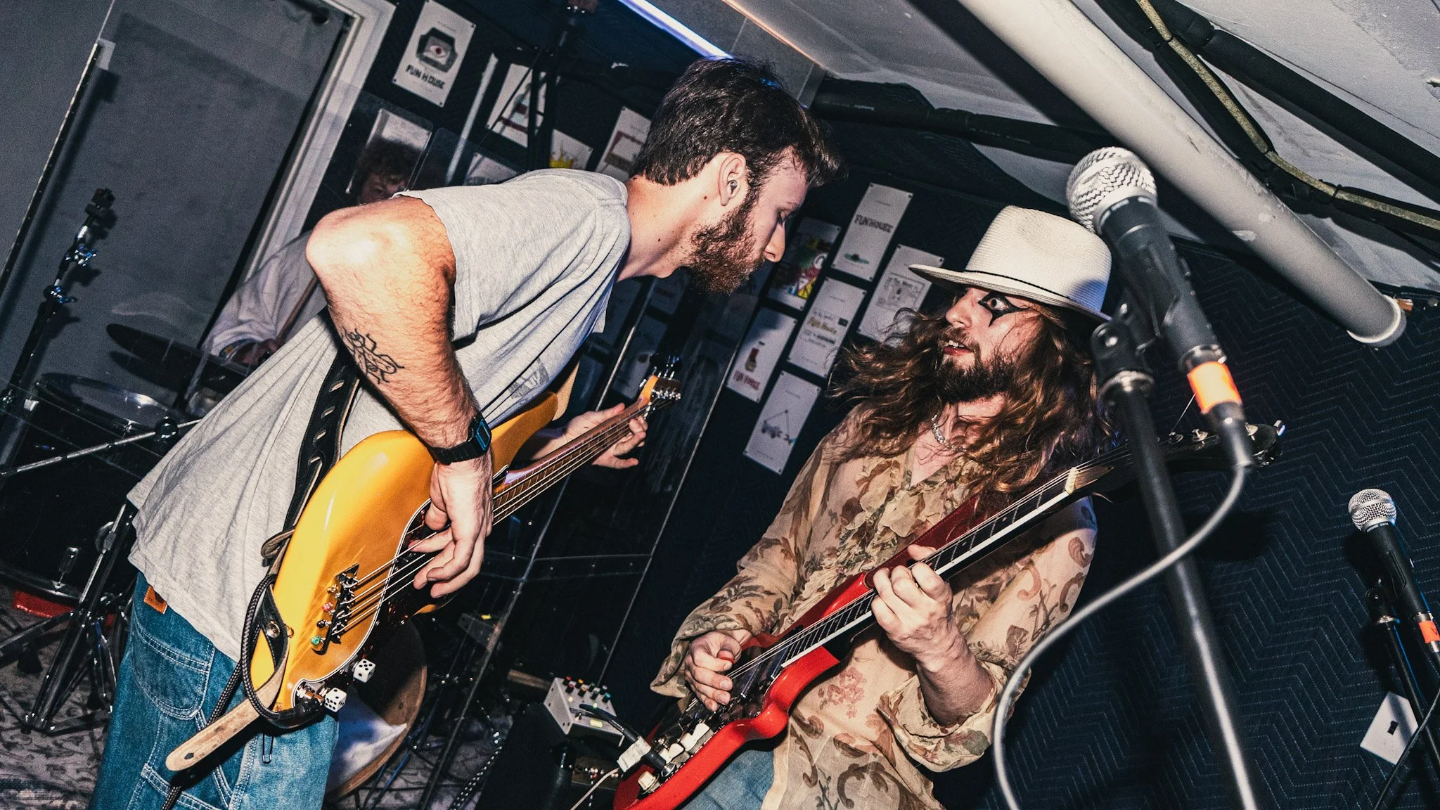 Two men playing electric guitars in a recording studio, with one wearing a beige hat and the other with a beard and tattooed arm, microphone in the foreground, and music studio posters on the wall.