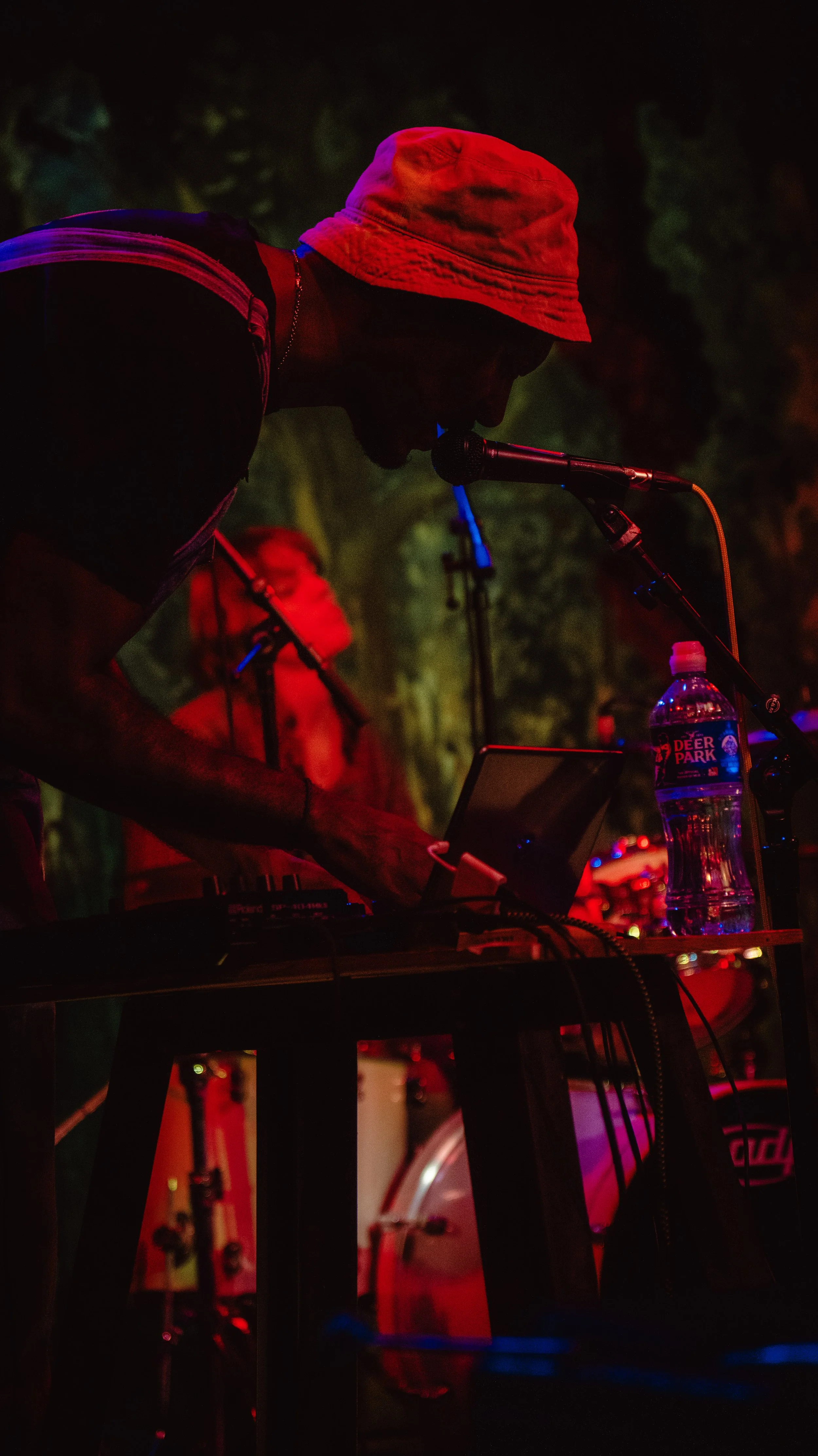 Person wearing a red bucket hat and dark clothing performing with electronic music equipment at night, illuminated by red and blue lights, with a water bottle on the table.