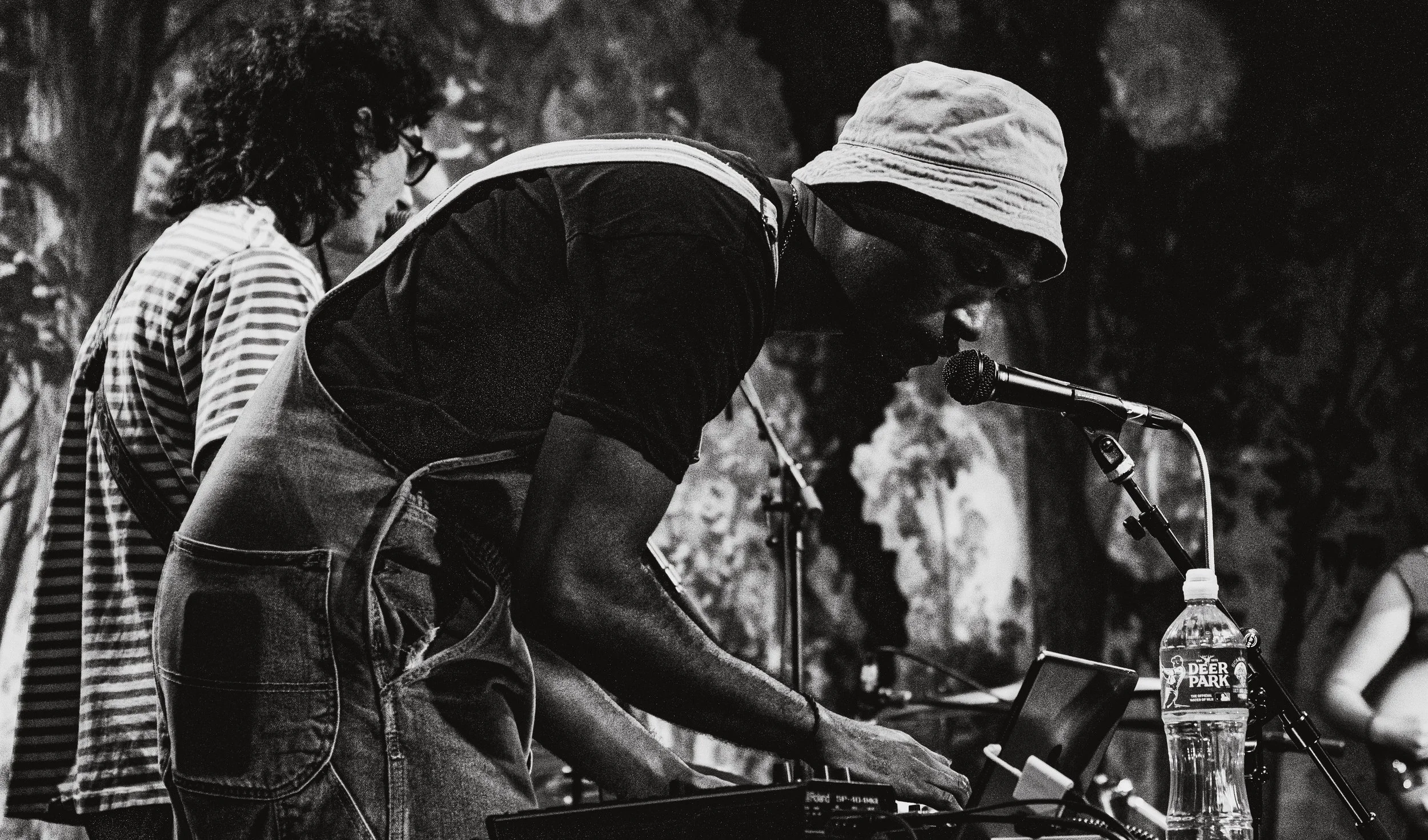 Black and white photo of a person in a hoodie and sunglasses speaking into a microphone at an outdoor event, with other people visible in the background.