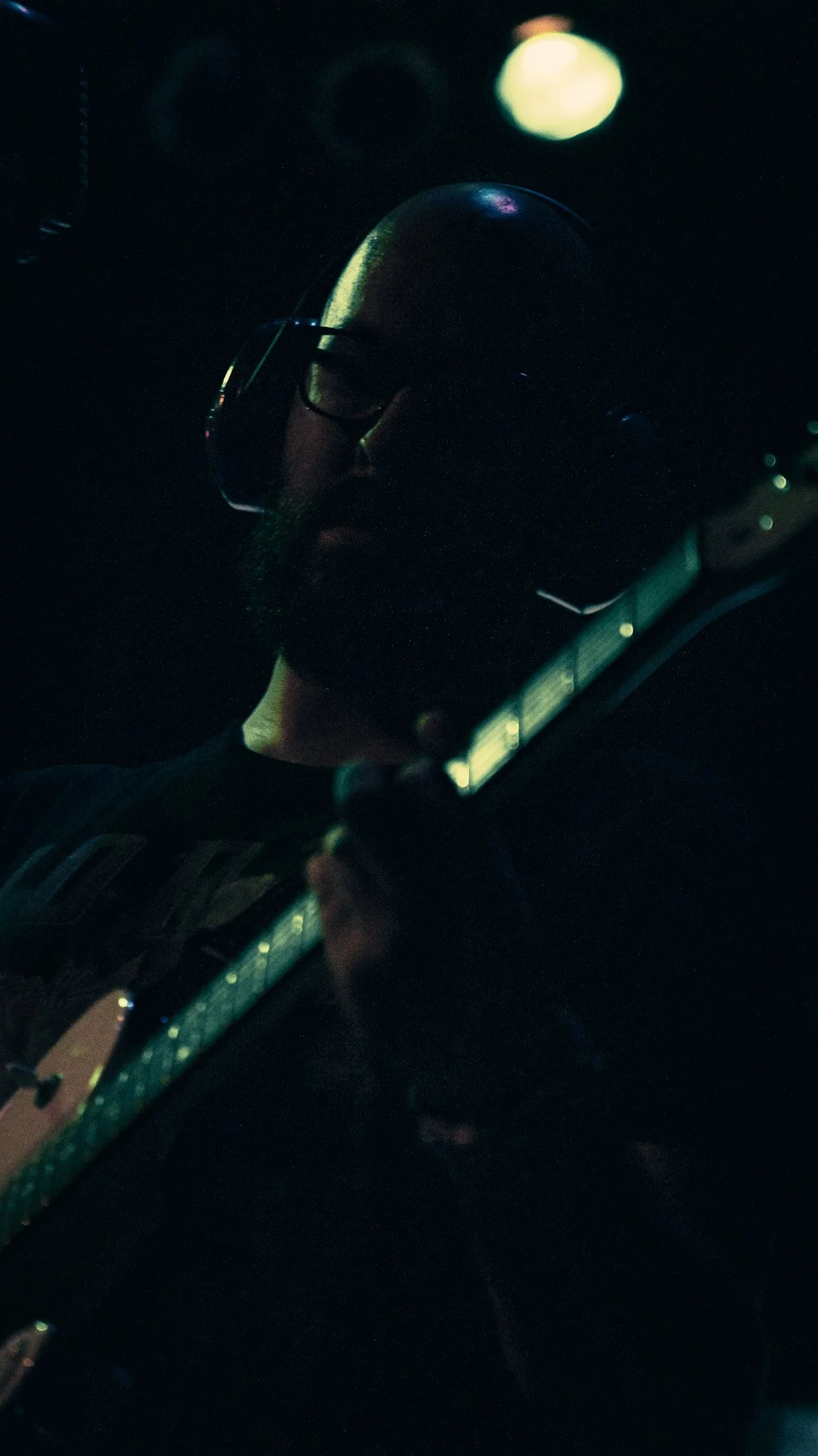 A person wearing glasses plays an electric guitar in a dark setting, illuminated by a faint light source.