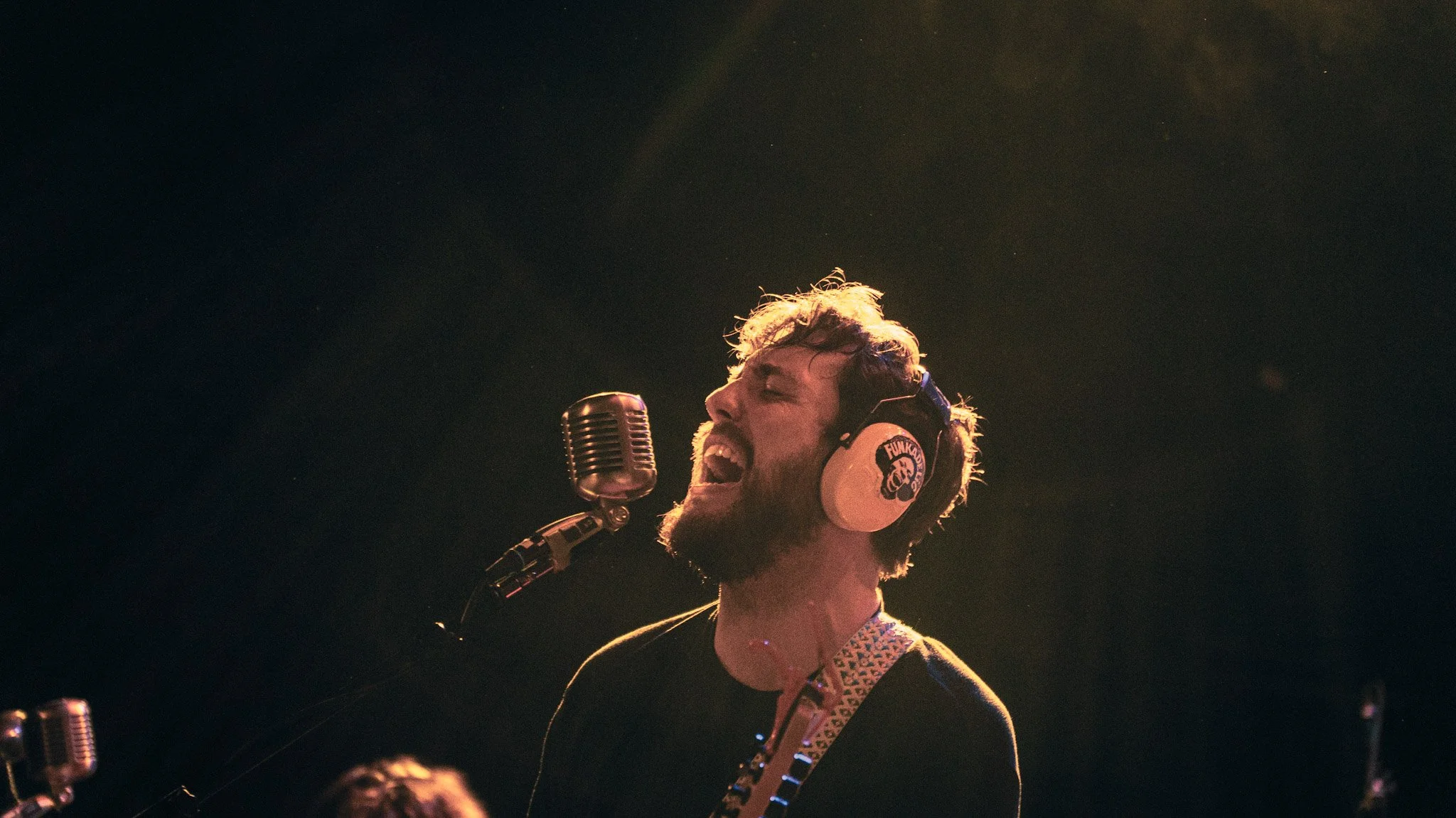 A man singing passionately into a microphone while wearing headphones, against a dark background with a faint depiction of stars or lights.