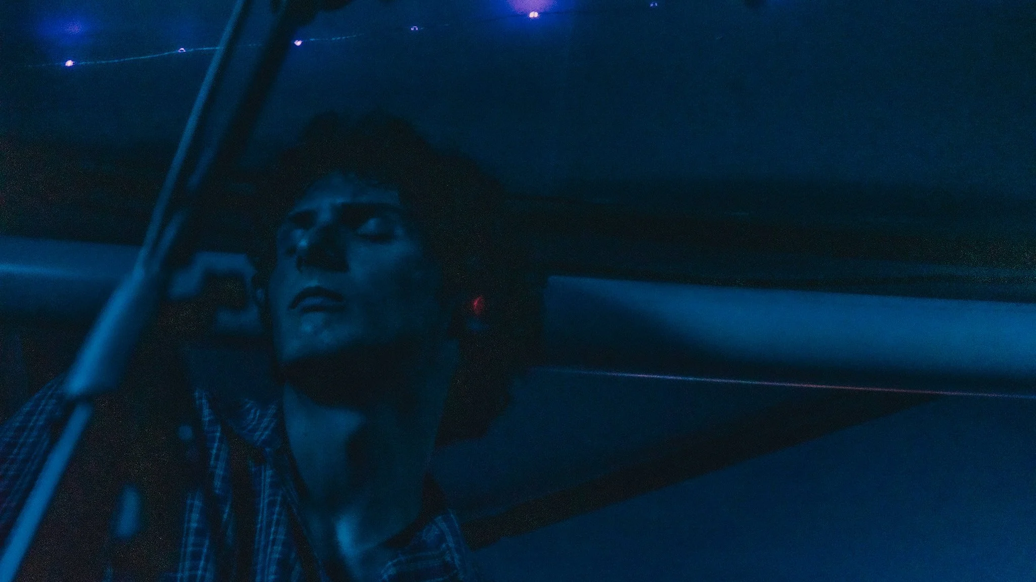 A person with long hair in a checkered shirt inside a vehicle at night, with visible dashboard and a dark background.