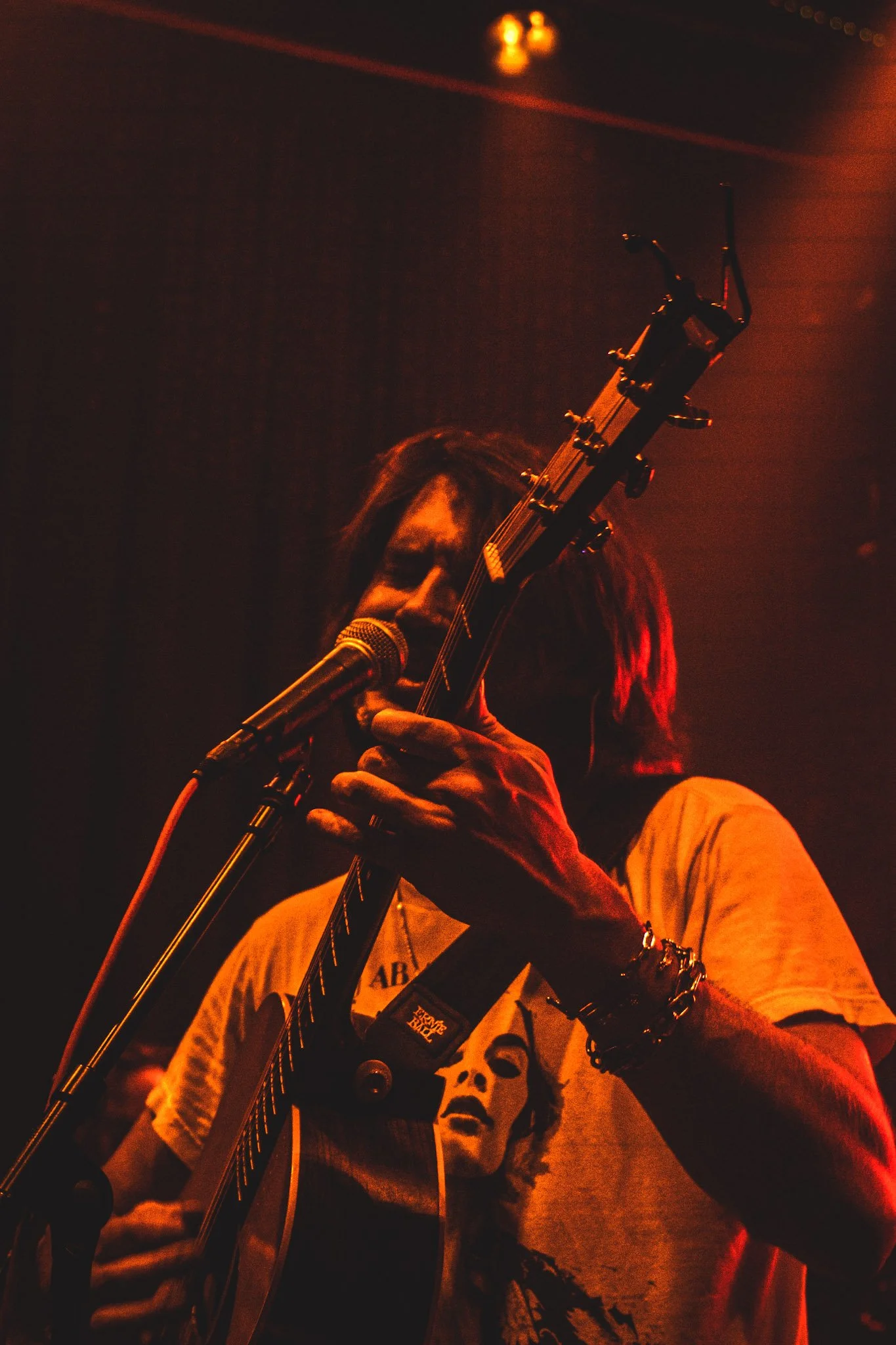 Musician with dreadlocks playing guitar and singing into a microphone on stage, dimly lit with red and orange tones.