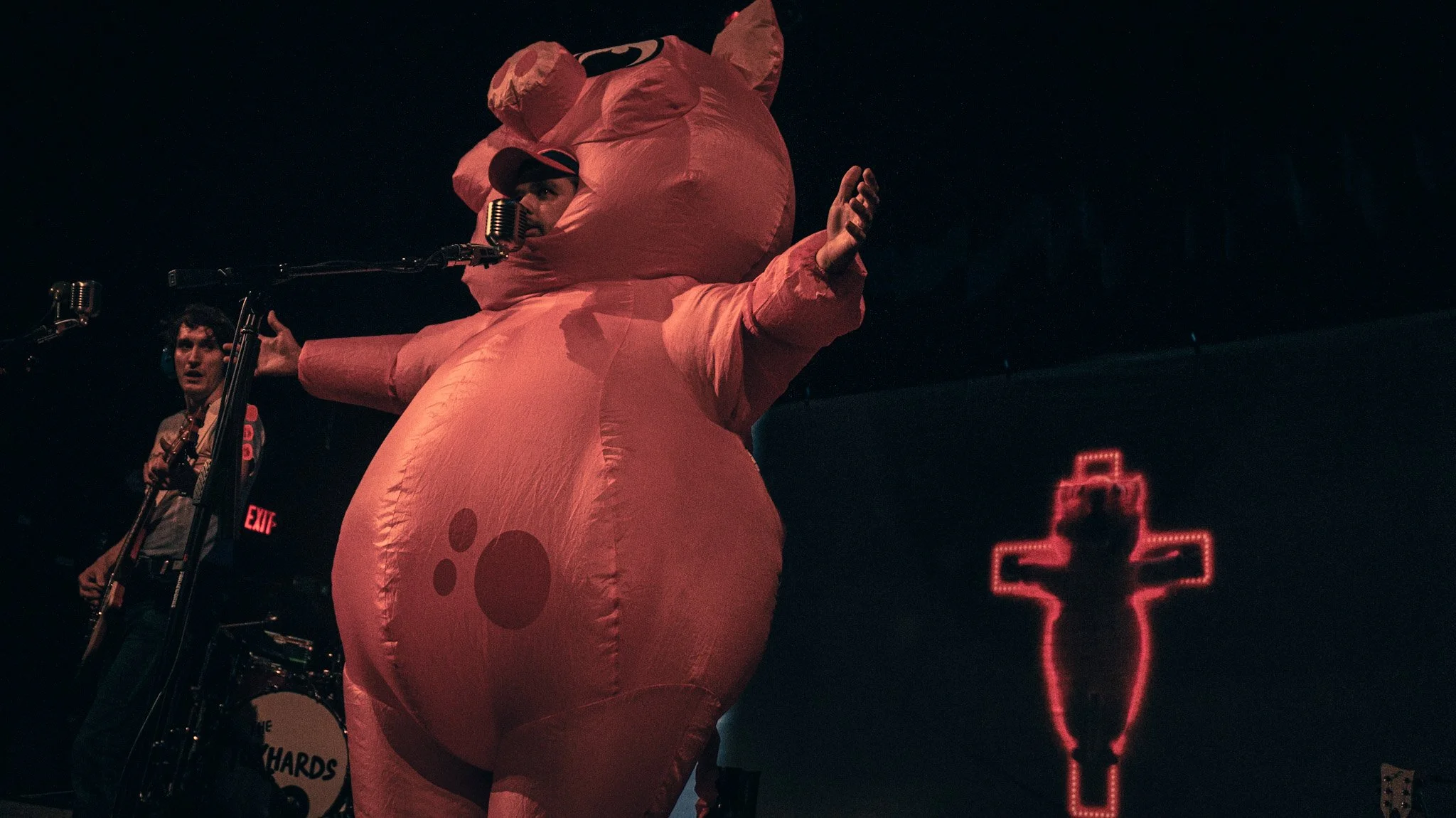 A person dressed in a large pink animal costume, possibly a pig, on stage with a microphone. There is another person playing guitar nearby, and a neon red cross symbol in the background.