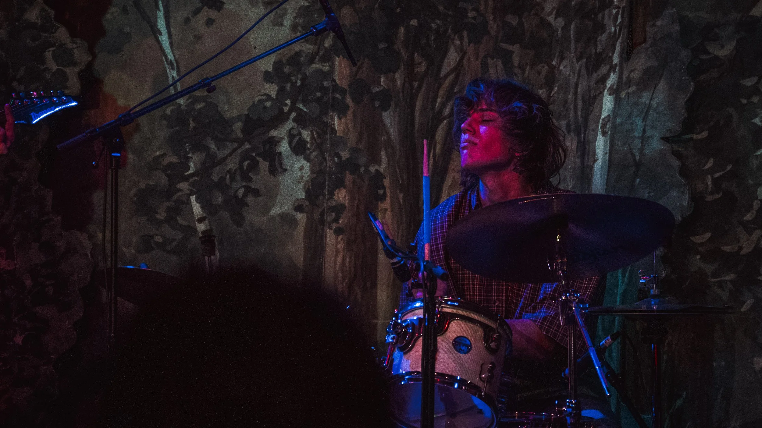 A woman playing drums in a dimly lit room with dark textured walls, illuminated by colorful stage lights.