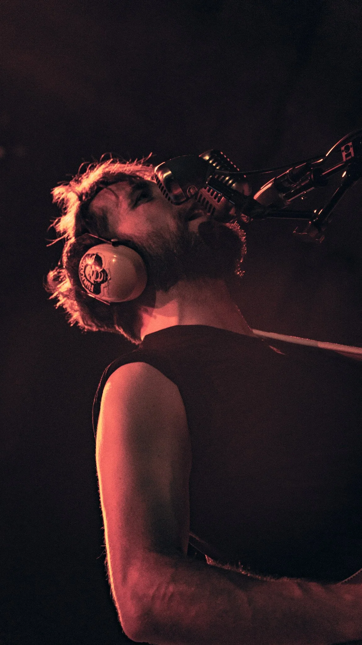 A man with a beard and messy hair wearing headphones and a black sleeveless shirt is singing into a professional microphone in a dark setting with red lighting.