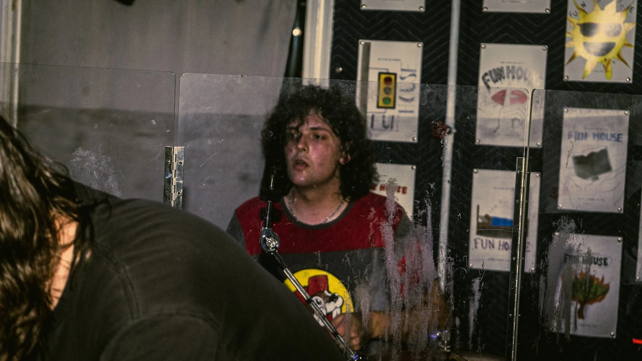 Young man with curly hair behind a glass partition, looking to the side, in an indoor setting with signs and posters on the wall.