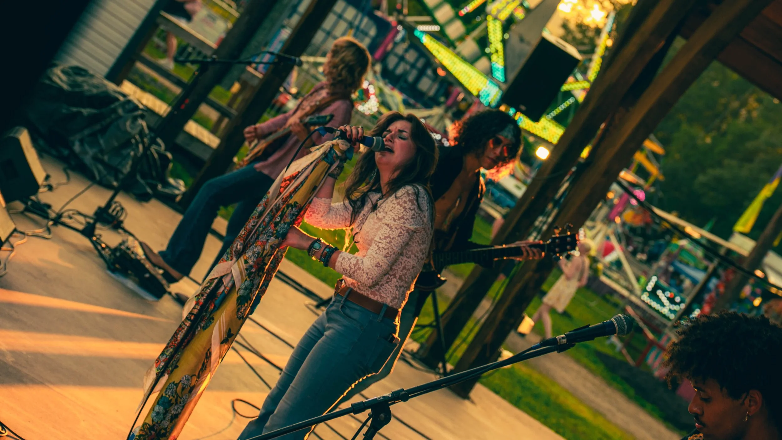 A group of musicians performing at an outdoor carnival or fair during sunset, with a singer holding a microphone in front of a guitarist, surrounded by colorful lights and carnival rides.
