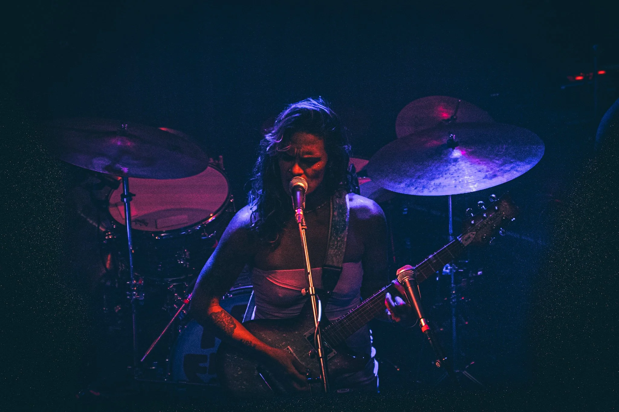 A female musician playing an electric guitar and singing into a microphone on a dimly lit stage with purple and blue lighting, surrounded by a drum set.