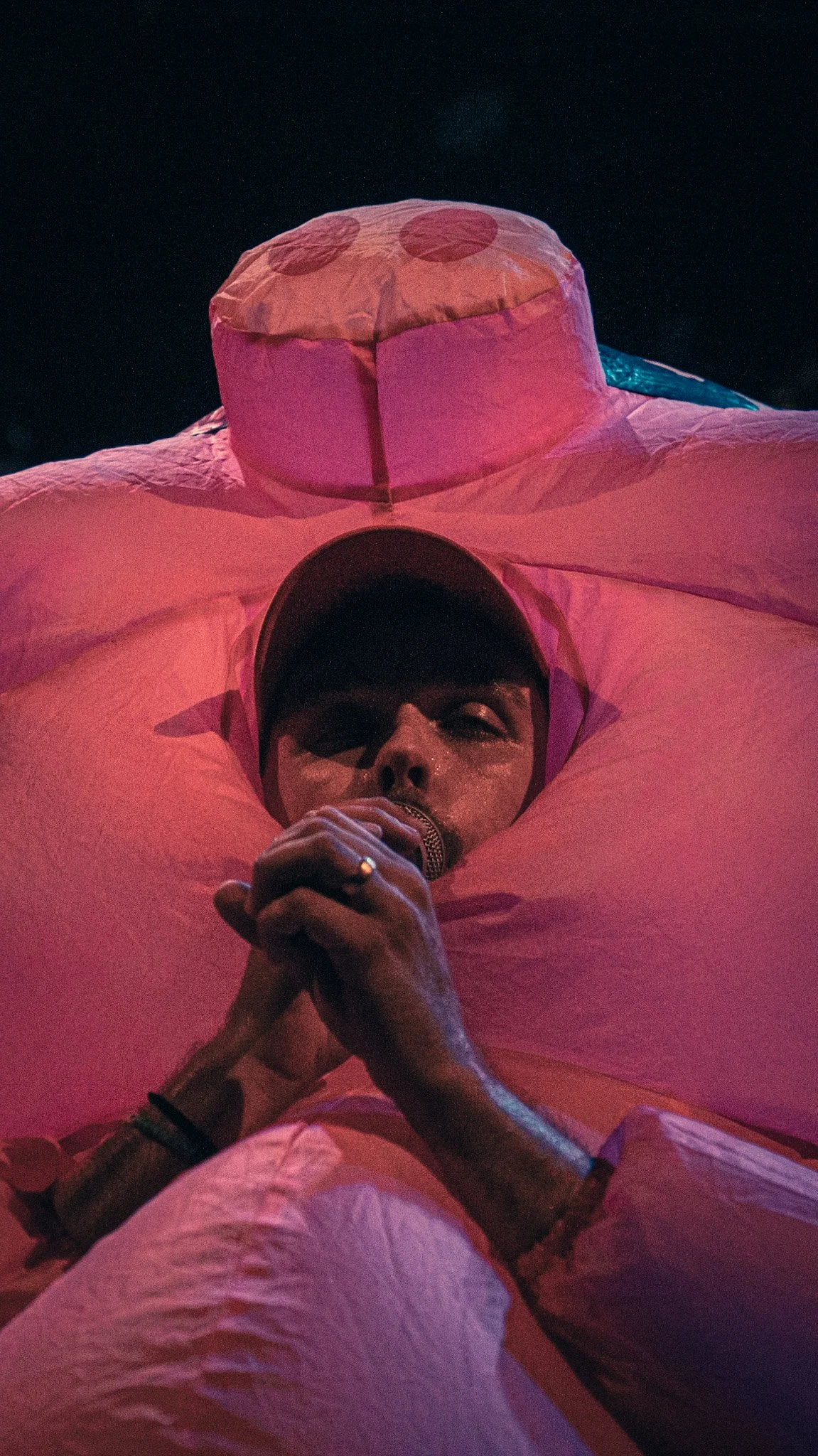 A person in a cap is lying down and holding a microphone near their mouth. The person appears to be singing or speaking, and the background is dark with dim lighting.