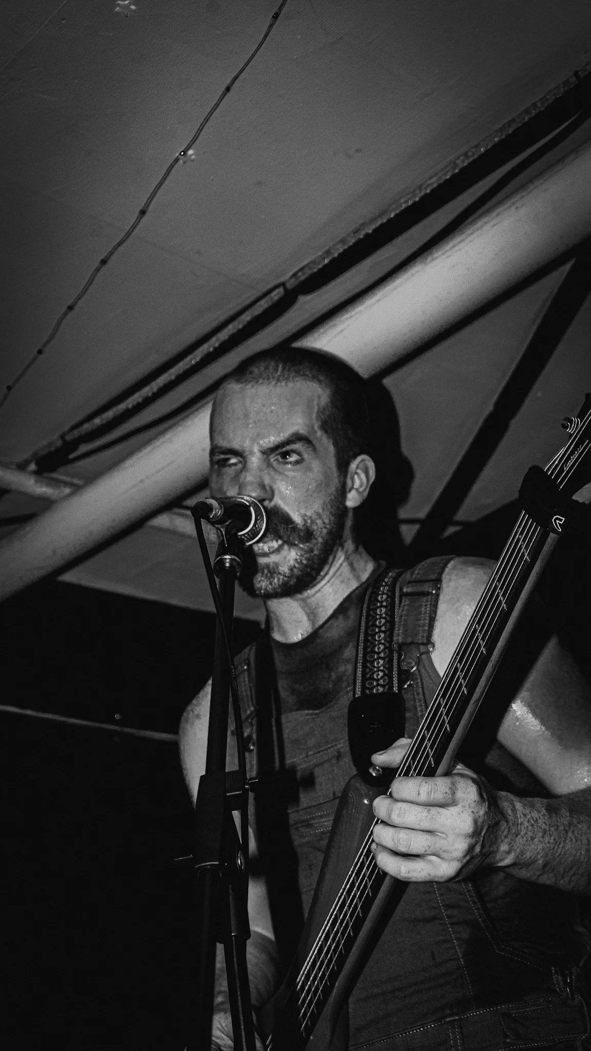 Black and white photo of a man with a beard singing into a microphone while playing an electric bass guitar, wearing a sleeveless shirt.