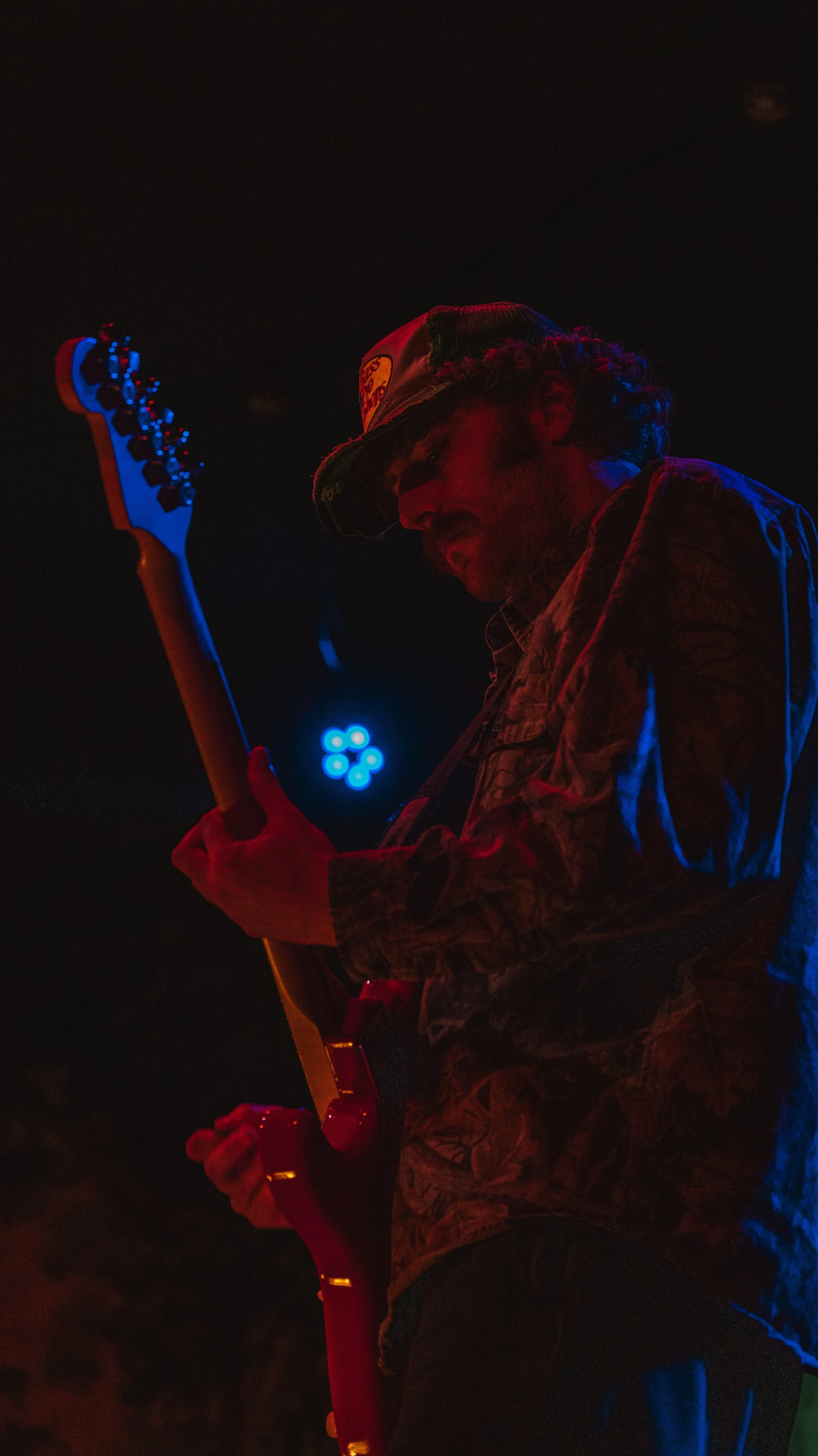 A musician playing an electric guitar on stage, illuminated by red and blue stage lights, with a dark background.