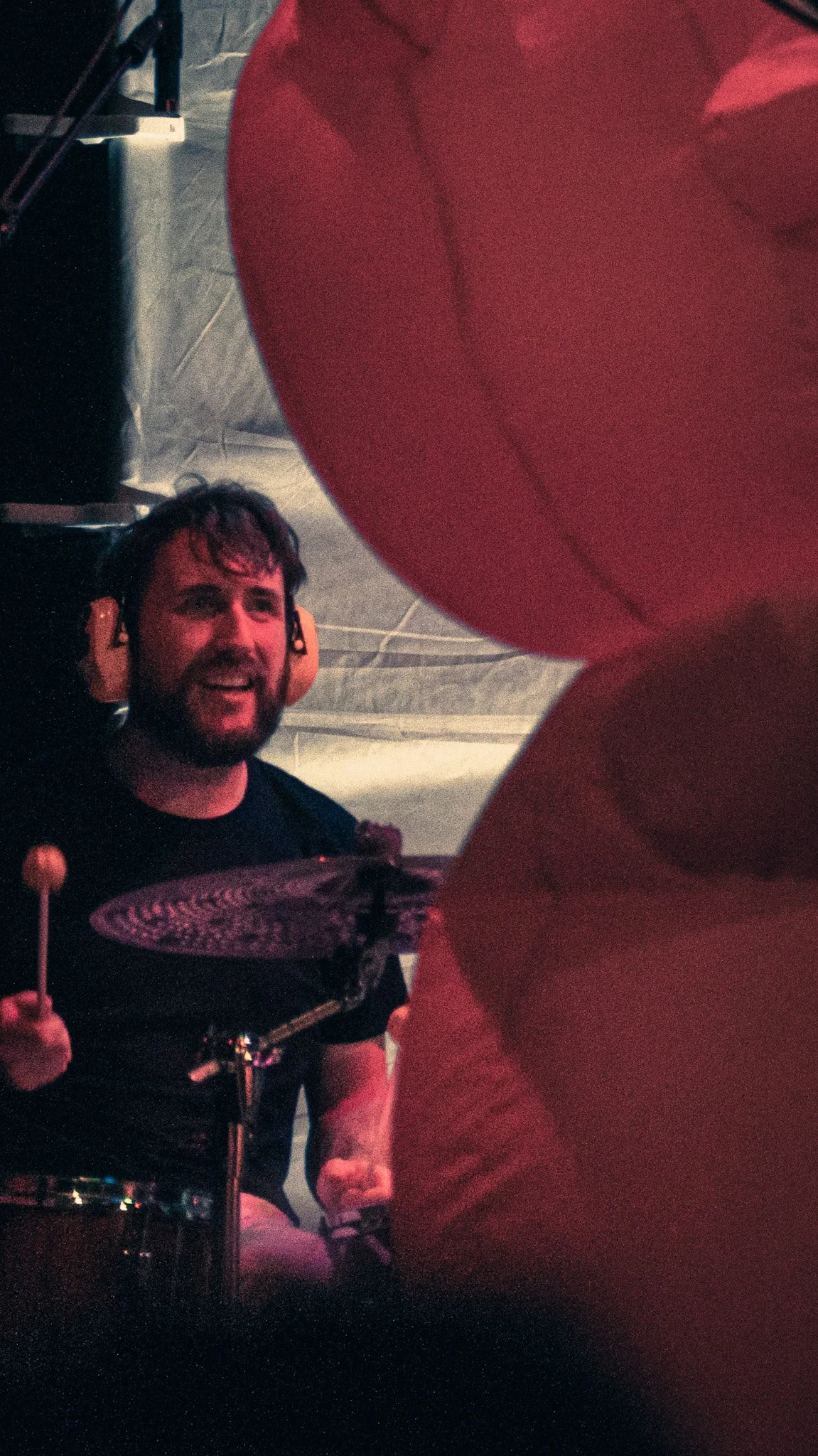 A man with a beard and glasses wearing headphones playing drums in a dimly lit setting, with a red object in the foreground partially obscuring the view.