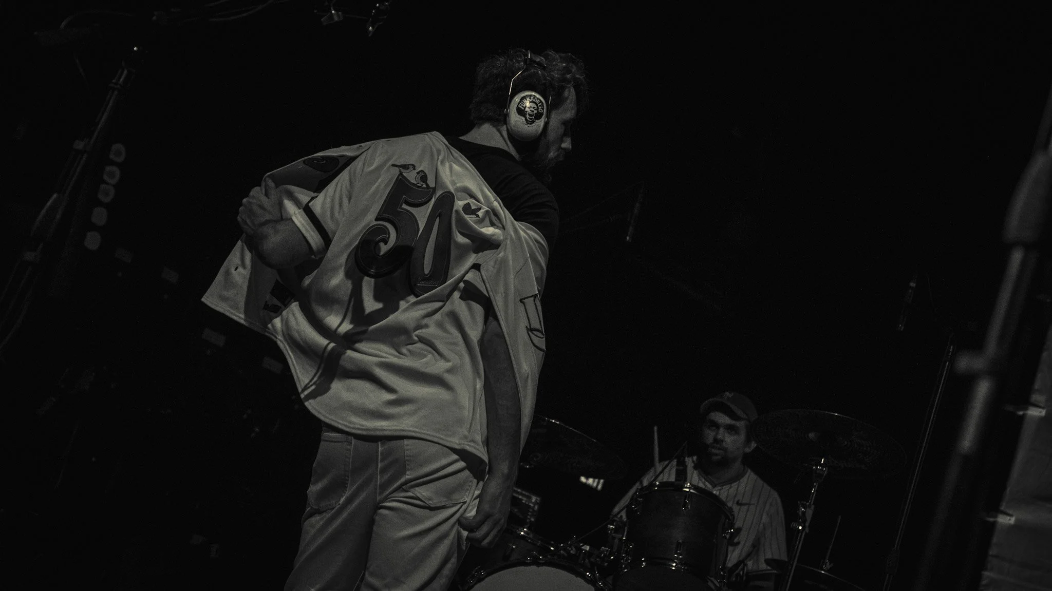 A man wearing a sports jersey with the number 50 and an emblem on the sleeve, listening to headphones, appears to be in a music studio or rehearsal space, with a person in the background playing drums.